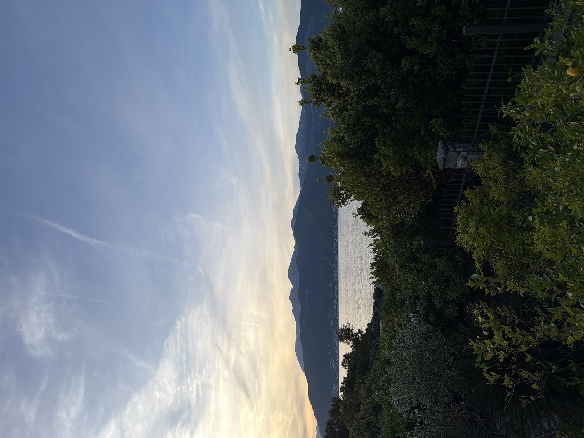 The image depicts a landscape scene in Gošići, Montenegro. The sky is blue with wispy clouds and contrails. A mountain range dominates the center of the frame. The sea is visible, reflecting sunlight. Lush green trees and foliage are in the foreground, obscuring parts of the mountain and sea. A dark fence and stone structure are visible among the vegetation.