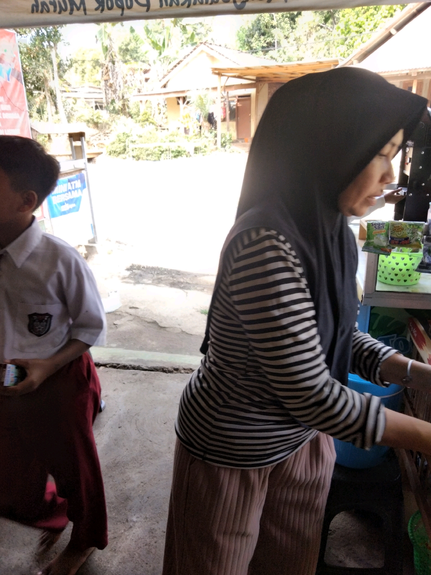 The image depicts an outdoor scene at what appears to be a small roadside kiosk in Sidorejo, Indonesia. In the foreground, a young person wearing a white school uniform shirt with a crest on the left chest and maroon pants is partially visible on the left side of the frame. They are holding a small, colorful object, possibly a phone or a pack of cigarettes. To the right, a woman wearing a black hijab and a black and white striped long-sleeved shirt and light pink ribbed pants is standing. She is looking down and to her right, potentially interacting with a customer or tending to the kiosk. Her right hand, adorned with a silver bracelet, is visible reaching towards the counter.
The kiosk itself has shelves displaying various products, including what appear to be packaged snacks or food items. A green basket with heart cutouts is visible on one of the shelves. The background shows a building with a light-colored roof and wooden beams, suggesting a simple structure typical of such stalls. Lush green foliage and trees are visible behind the building, indicating a rural or semi-rural environment. The ground surface is a rough, paved area. The overall impression is of a casual, everyday transaction taking place in a warm, sunny climate.