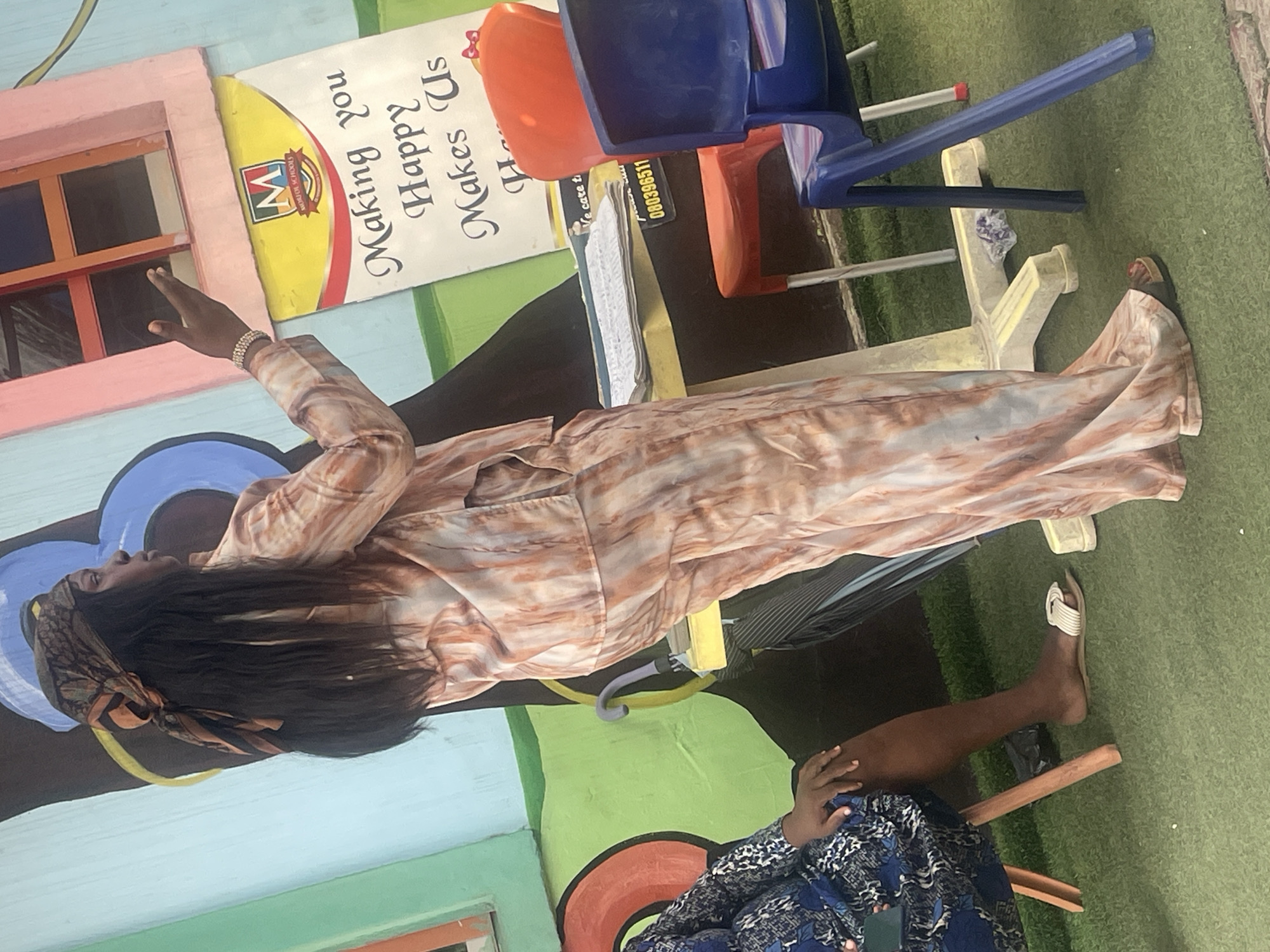 In this image, a person is sitting on a wooden stool in an outdoor setting, likely a schoolyard given the colorful murals and play equipment. The person is wearing a patterned dress and headscarf, with their right arm resting on their lap. Their left hand is raised, as if reaching for something out of frame. A blue plastic chair and an orange chair are visible behind them, as well as a table with some papers on it.
The scene appears to be in Abuja, Nigeria. The murals suggest a vibrant and child-friendly environment, with bright colors and playful imagery. The text on one of the murals, though partially obscured, reads "Making you Happy Makes Us". The time of day is difficult to determine definitively, but the lighting suggests it might be daytime. The overall mood of the image is relaxed and casual.
Another person's legs and feet, wearing sandals, are visible in the lower right portion of the image, indicating the presence of at least two people in the scene. The ground is covered in green astroturf, typical of many outdoor recreational areas. The image is oriented vertically, with the top of the frame showing a pink and yellow structure, possibly part of the building.