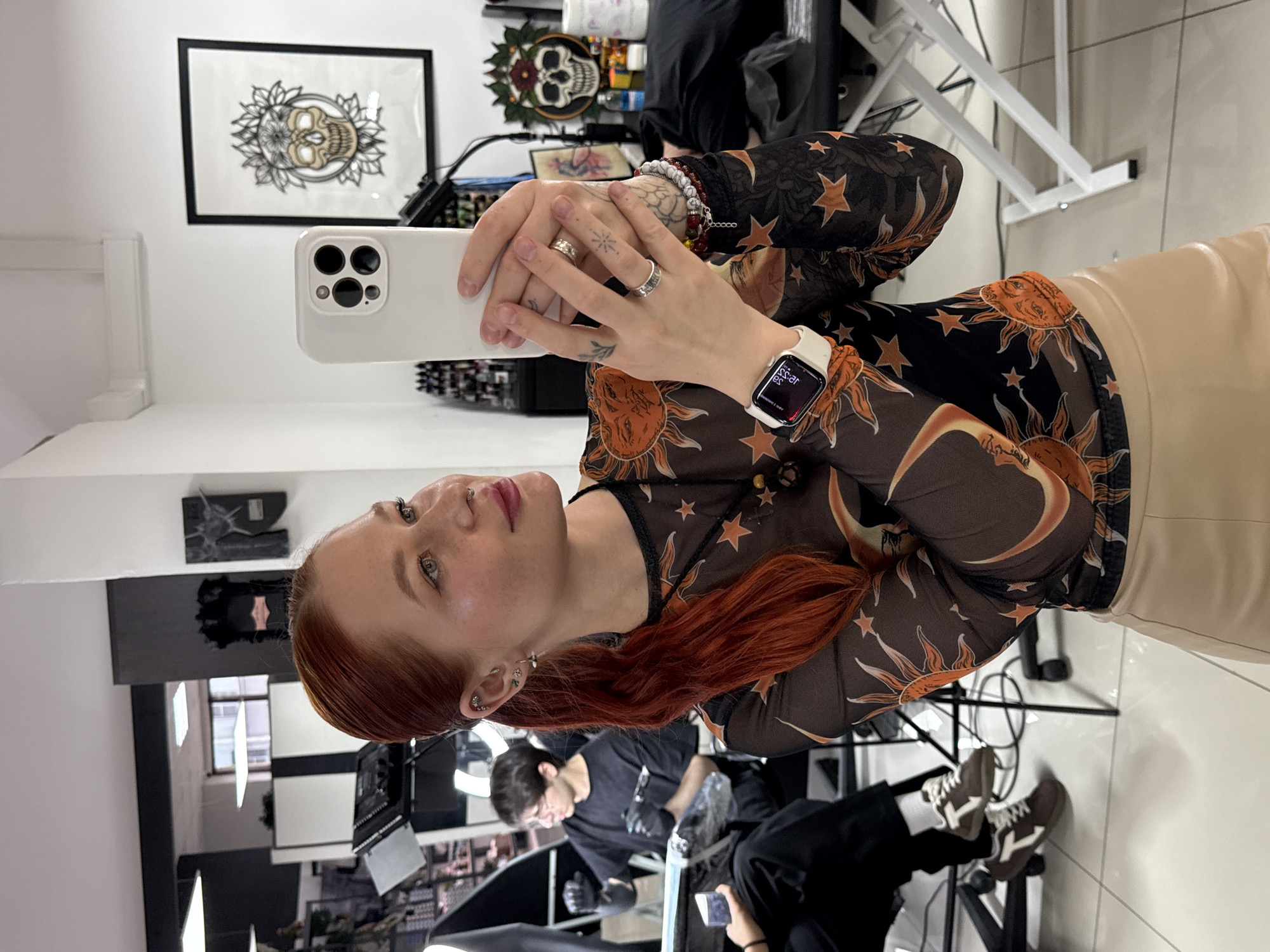A young woman with red hair is taking a selfie with a white smartphone in a room that appears to be a tattoo studio. She is wearing a black top with a sun and star pattern and a light beige garment. She is also wearing an Apple watch. In the background, a person in black attire is working, and tattooing equipment is visible. Artwork, including a skull illustration, adorns the walls. A black-and-white themed workstation is visible near the back of the room. The location is Rostov-on-Don, Russia.