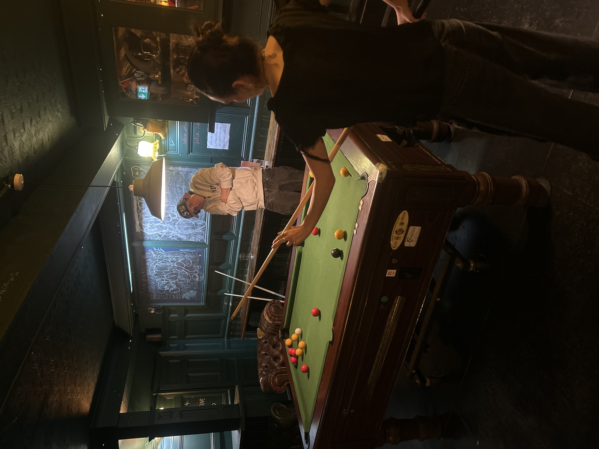 The image depicts a billiards game in progress inside a pub, likely in London, United Kingdom. Two individuals are present: one, in the foreground, is bent over the table, cue in hand, poised to make a shot; the other stands nearby, watching. The billiards table is green, with several balls scattered across its surface. The pub interior features dark wood paneling, chalkboard-style wall art, and overhead lighting, creating a dimly lit, traditional atmosphere.