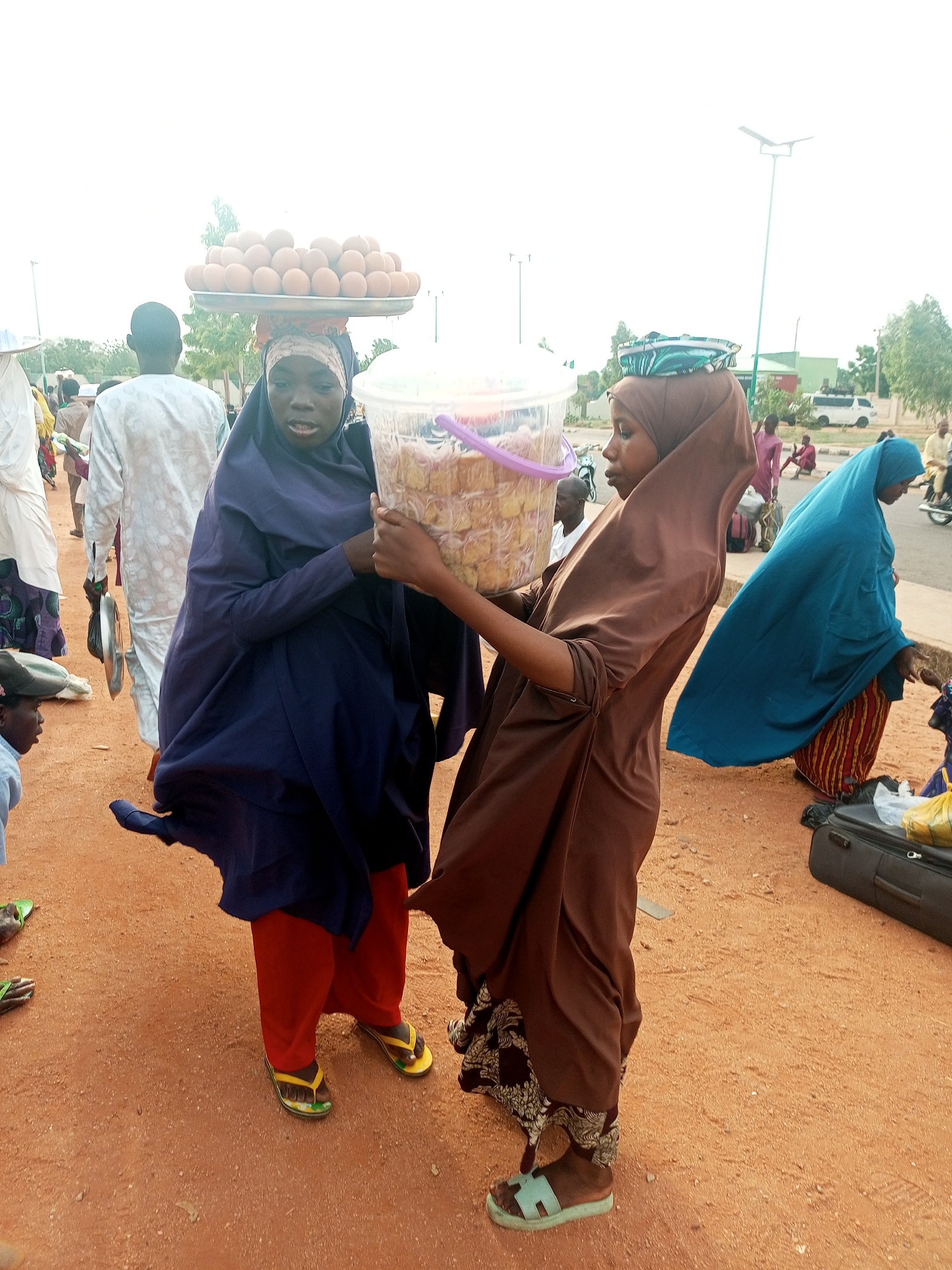The image depicts two women in Gombe, Nigeria, one carrying a tray of eggs on her head and the other holding a clear plastic bucket containing food items. The woman carrying the eggs wears a dark blue robe over red pants and stands to the left of the frame. The second woman, wearing a brown robe, is partially supporting the bucket with her hands. Both women wear head coverings typical of Muslim dress. They are standing on a dirt ground with other people and objects in the background, including suitcases, other pedestrians, and vehicles along the street. A blue-robed woman stands closer to the street. Streetlights are visible in the background indicating an urban or semi-urban setting.