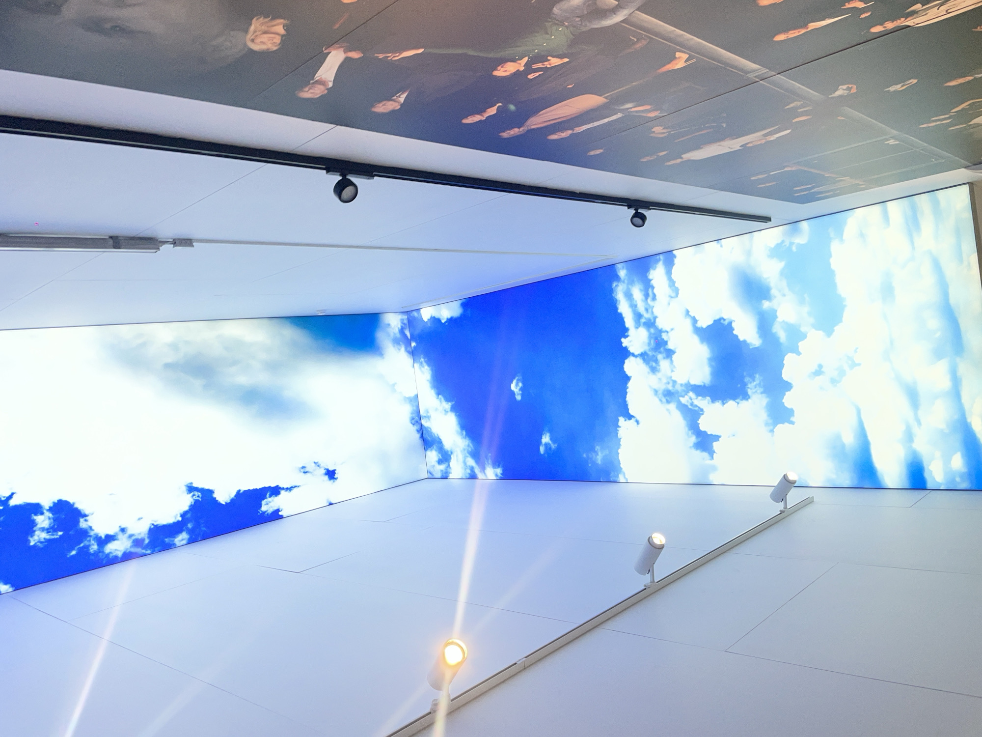 The image shows an interior space, possibly a room or hallway, in Khimki, Russia. The scene has multiple ceiling and wall displays.
The upper ceiling displays a dark-toned image of a group of people, seemingly upside down from the viewer's perspective.
Below this, there's a section with a white ceiling and a dark, track lighting system.
The walls display an image of a blue sky with white clouds. On the lower right wall, there's a lighting fixture with three adjustable spotlight-style lamps. The lighting is directed upwards towards the "sky" display. The walls are white with a grid-like pattern.