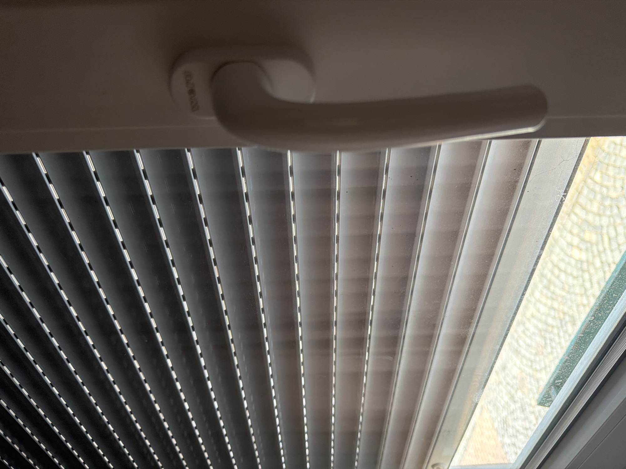 The image shows a close-up of a window with closed horizontal blinds. The blinds are dark gray with thin, lighter-colored horizontal lines running across them, giving them a textured appearance. To the right of the blinds is a white window frame with a white window handle. The handle is curved and in the down position, suggesting the window is closed. The bottom of the image shows a glimpse of the exterior through the blinds, revealing a paved area with a circular pattern and some greenery. The lighting suggests it is daytime, but the blinds are closed, obscuring most of the external view. The overall scene is indoors, looking out.