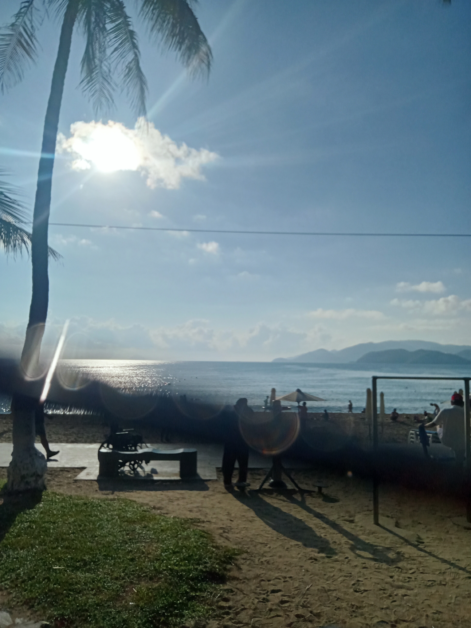 The image depicts a beach scene in Nha Trang Ward, Vietnam, under a bright, sunny sky with scattered clouds. A tall palm tree stands on the left side of the frame. The ocean is visible, with sunlight reflecting off the water. In the middle ground, there are people on the beach, including at least one person standing near what appears to be a bench. To the right, there is a structure with bars and figures of people on the beach. In the foreground, there is green vegetation and sandy ground.