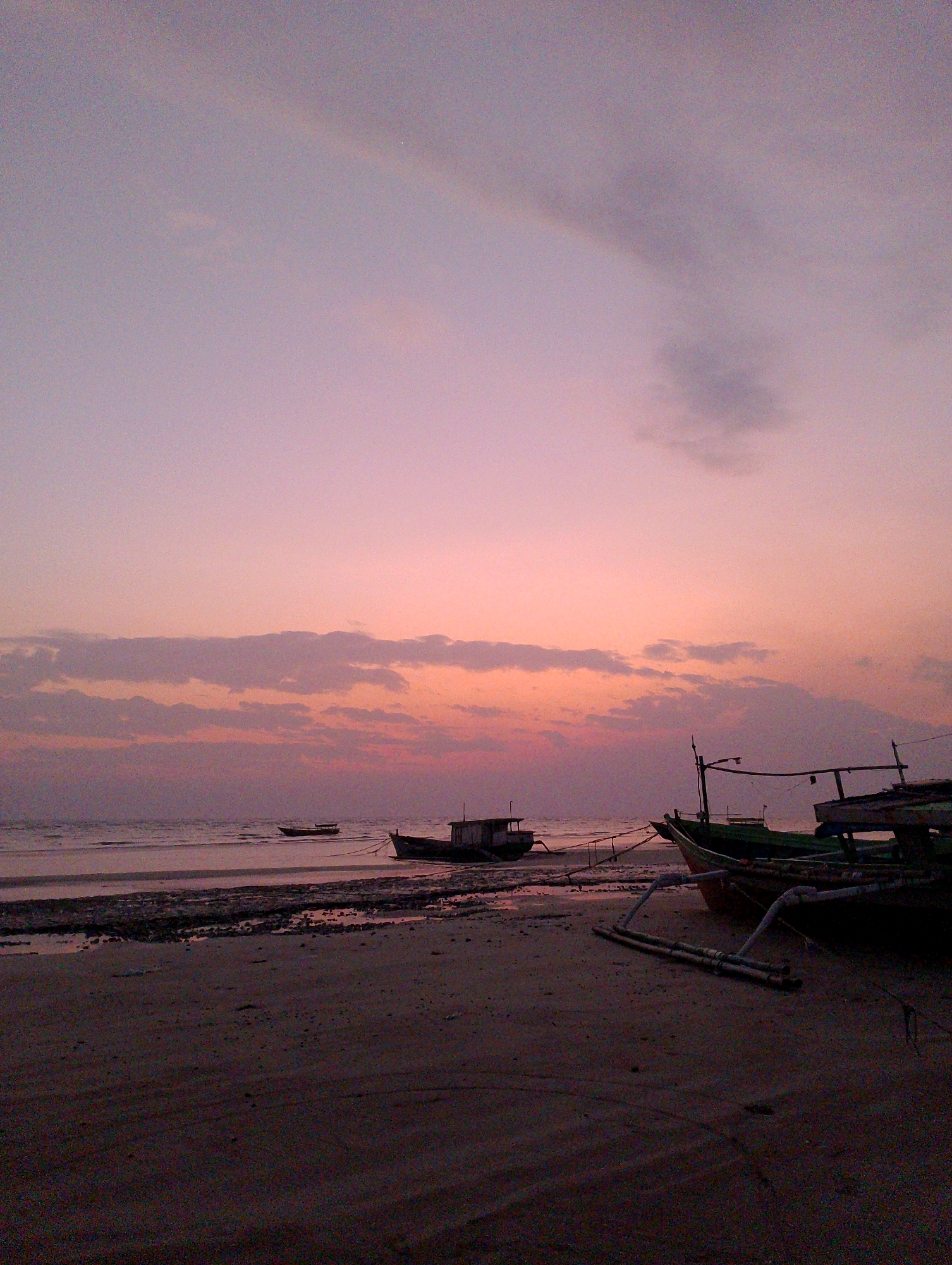 This is a photograph of a beach at dusk, likely in Merauke, Indonesia. The sky is filled with soft hues of pink and purple, transitioning to a pale lavender at the top. Scattered, wispy clouds are visible, catching the last light of the setting sun.
In the foreground, a sandy beach is visible, with a dark, wet patch towards the left, indicating the receding tide. Several fishing boats are anchored or beached on the sand. In the middle ground, two smaller boats are moored in the shallow water. Further to the right, larger fishing vessels are pulled up onto the beach, their dark hulls contrasting with the soft sky. Some of the boats have visible structures, such as masts and railings, and ropes are used to secure them. The water's surface is calm and reflects the sky's colors. The overall atmosphere is tranquil and serene, typical of a coastal scene at twilight.