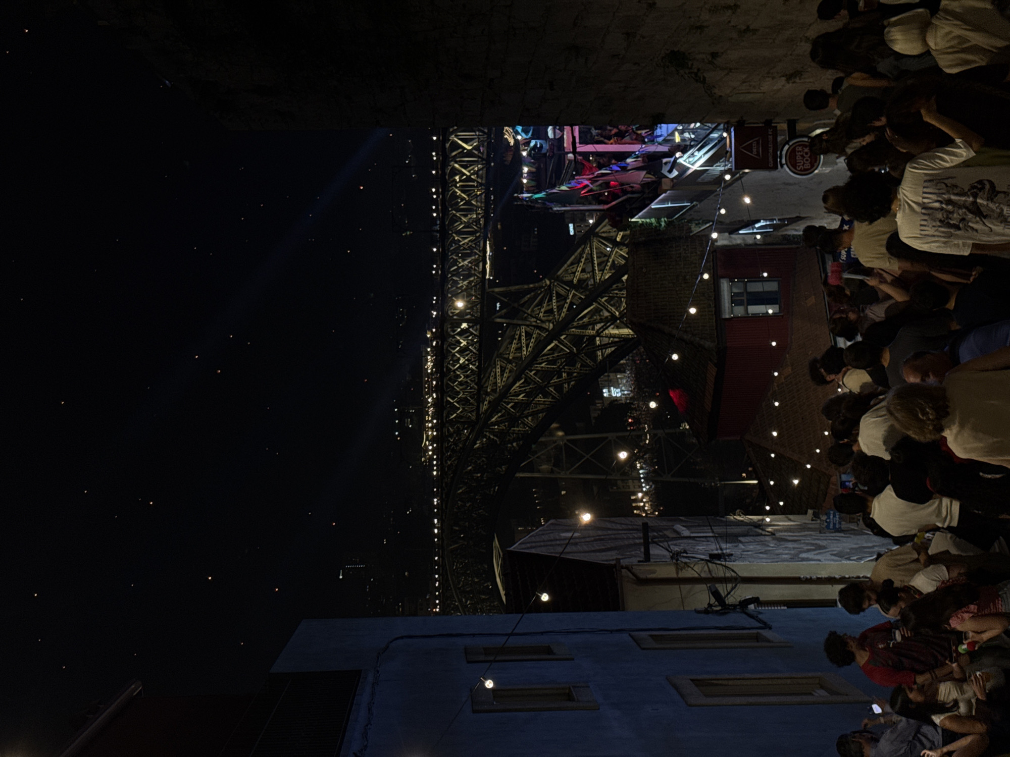 The image depicts a bustling outdoor gathering at night, likely a festival or a social event. A large, illuminated bridge, possibly the Dom Luís I Bridge in Porto, Portugal, dominates the background, its intricate metalwork and strings of lights creating a dramatic backdrop against the dark sky. The sky is filled with numerous small, glowing orbs, suggesting either distant lights, fireworks, or possibly sky lanterns.
In the foreground and midground, a crowd of people is visible, gathered in what appears to be a street or an open area adjacent to buildings. They are casually dressed and seem to be mingling, talking, and enjoying the evening. The atmosphere appears festive, with strings of fairy lights strung across the scene, casting a warm glow. Some individuals are holding phones, possibly taking pictures or videos of the event.
There are visible signs with text. One sign reads "SUPER BOCK," a well-known Portuguese beer brand, and another partially visible sign appears to be a logo for "DELTA," likely referring to Delta Cafés. These brands suggest the location is likely in Portugal and the event may involve food and drink vendors. The buildings in the foreground are illuminated, showing architectural details like windows. The overall scene suggests a vibrant and lively nighttime celebration in an urban setting, with a mix of natural and man-made light sources contributing to the atmosphere. The time of day is clearly night, and the weather appears to be clear.