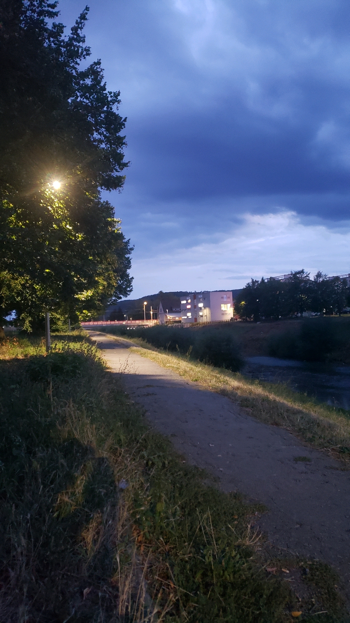 The image depicts a path along a riverbank in Prešov, Slovakia, at dusk. A large tree with illuminated leaves from a streetlamp stands on the left. The path, made of dirt and gravel, curves away into the distance. On the right side of the path, a river flows, its surface rippled and dark. Across the river, a white building complex with lit windows is visible, set against a backdrop of trees and a hill. The sky is a deep blue with dark, dramatic clouds, indicating twilight or approaching night. The overall scene suggests a serene, natural setting within an urban environment.