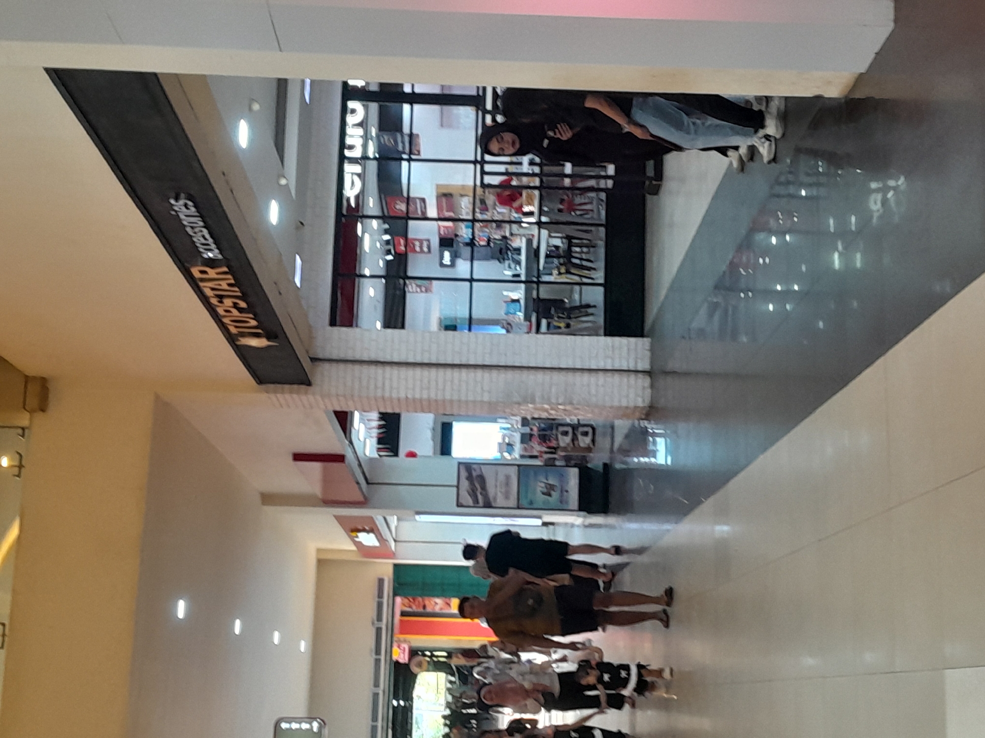 The image depicts a commercial walkway within a shopping complex, likely in Sempusari, Indonesia. The overhead lighting suggests an indoor environment. To the right, a store with the prominent signage "TOPSTAR accessories" is visible. The store's interior features large glass windows displaying merchandise. Several individuals are present in the walkway. A man in a tan t-shirt and shorts walks towards the camera, accompanied by a small child in a black and white outfit. Further into the walkway, other shoppers are visible, including a person in a black hijab standing near the entrance of a store. The floor is a highly reflective dark surface, creating mirror-like reflections of the surroundings and the lighting. The architecture suggests a modern shopping mall with tiled walls and suspended ceilings.
