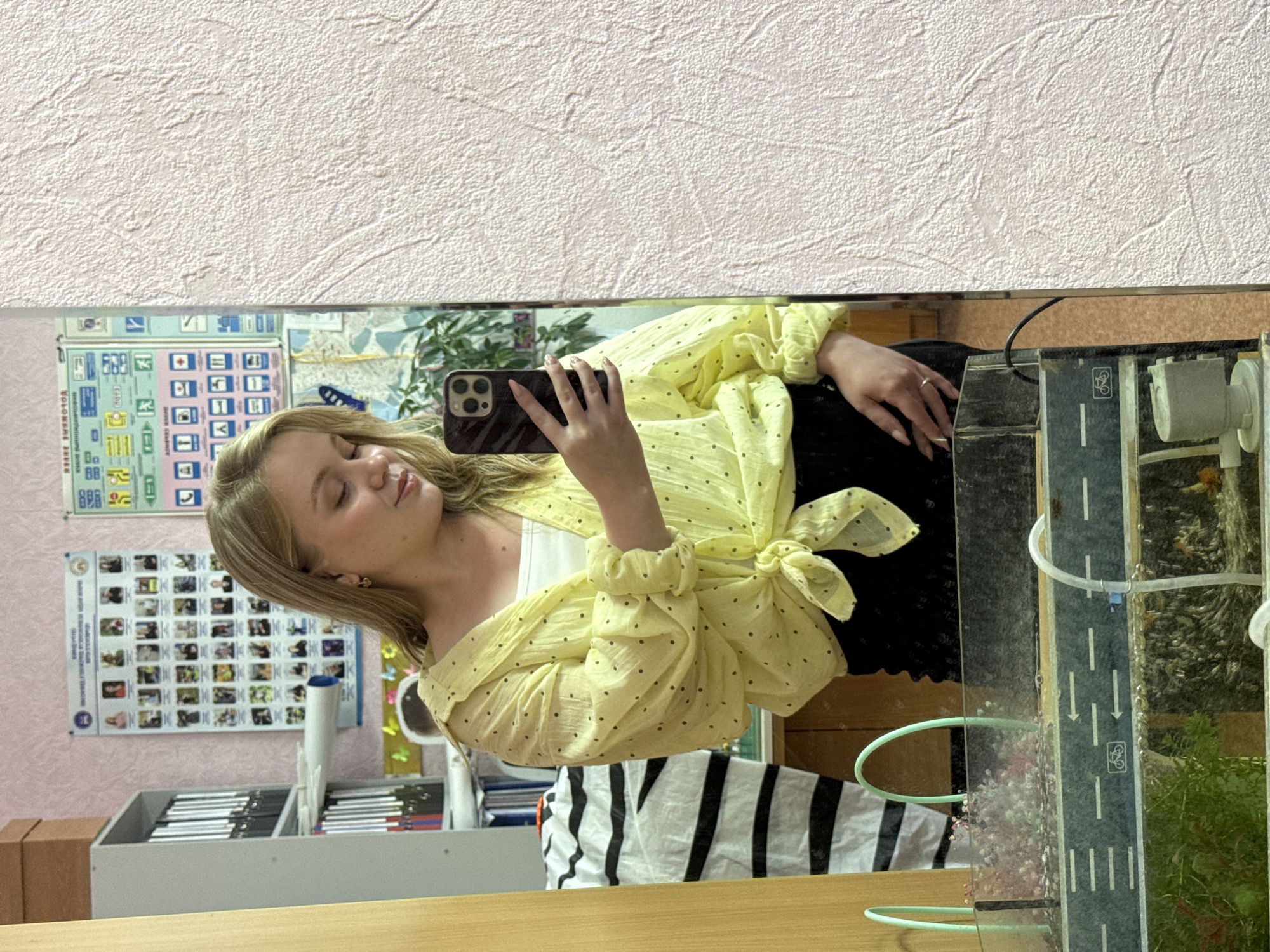 A young woman with blonde hair, wearing a yellow polka dot shirt and black pants, takes a mirror selfie in an indoor setting. She is holding a smartphone in her right hand. In the background, there are posters on the wall, bookshelves filled with binders, and a shelf with what appears to be a small aquarium. The overall environment suggests an office or educational space. The image is taken from the chest up.