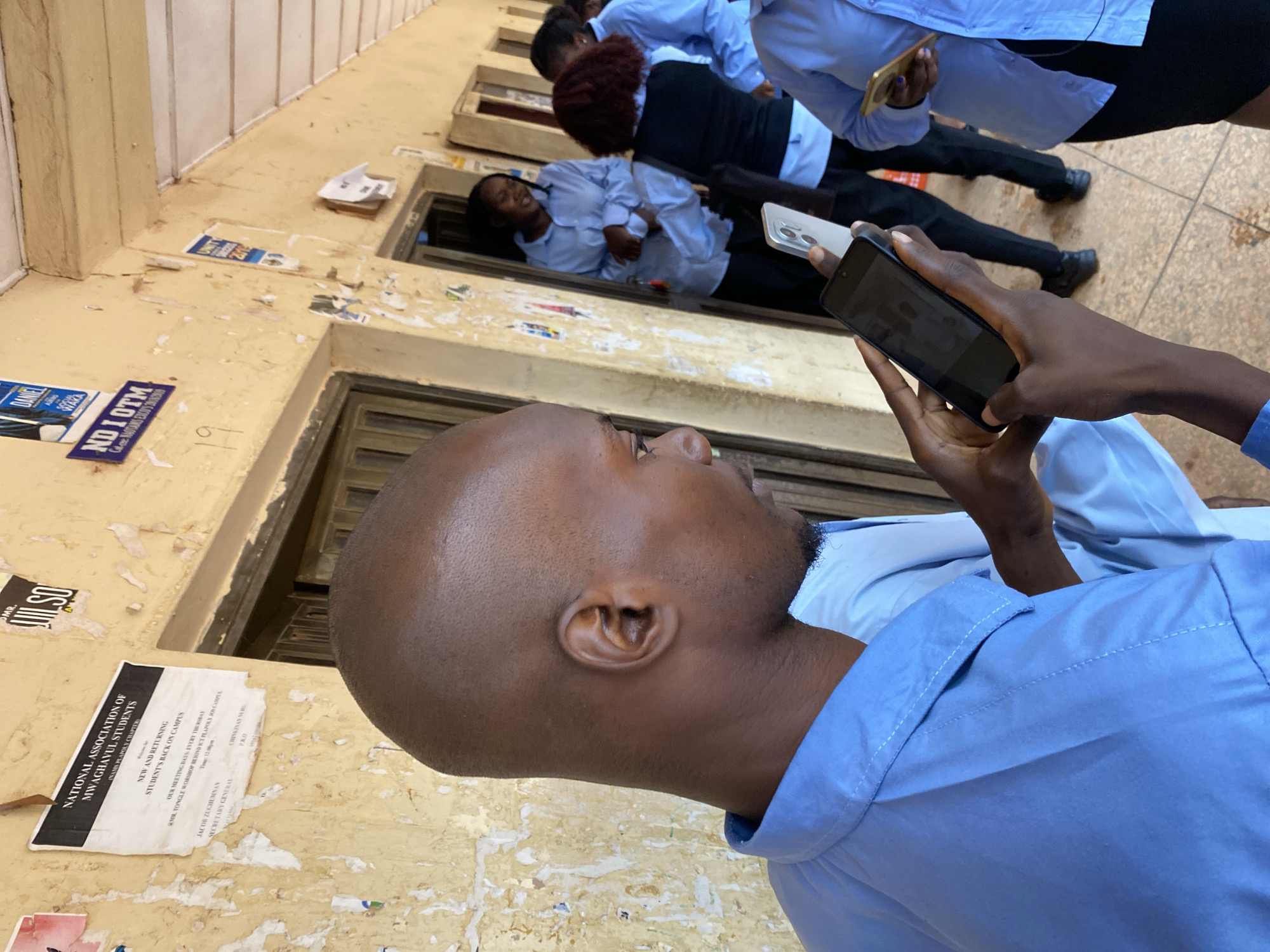 A photo, possibly taken in Jos, Nigeria, shows a person holding a smartphone, taking a picture of other people, some of whom are positioned in what appear to be recessed areas in a building's exterior wall. Most people are wearing light blue shirts, and one has a dark blazer. The exterior wall has posters and signs attached to it, with visible text and logos. One sign reads, "NATIONAL ASSOCIATION OF MWAGHAVUL STUDENTS." Additional, partially obscured signs are also visible. The scene suggests an outdoor location with architectural elements and informational posters displayed.