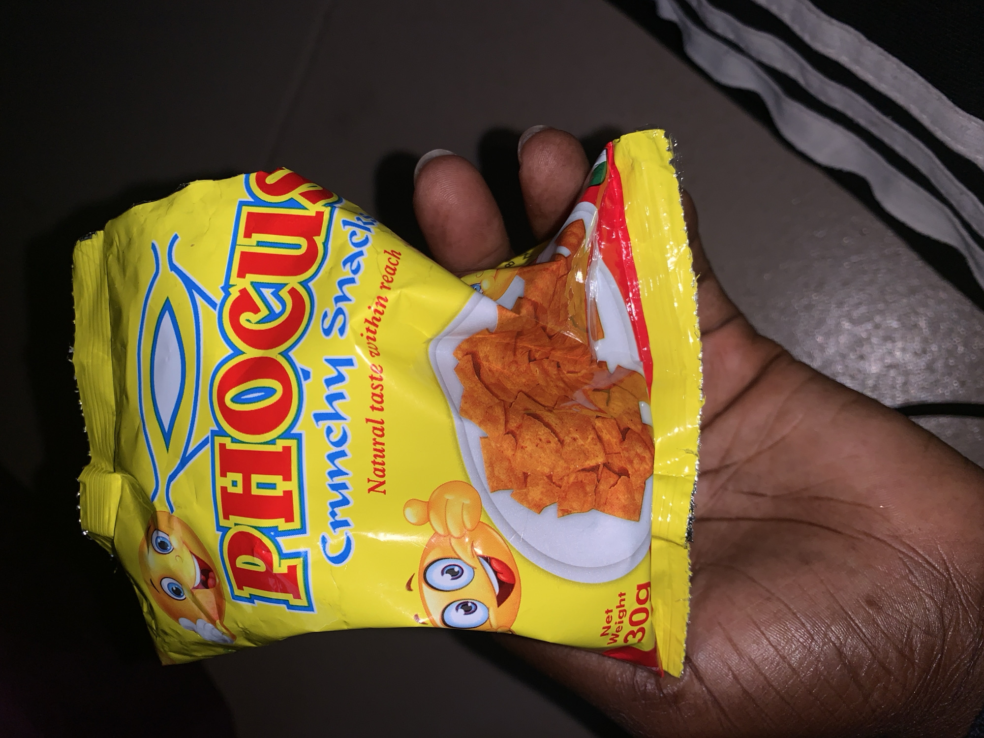 A person's hand is holding a yellow bag of "PHOCUS" brand crunchy snacks. The bag has a cartoon smiley face giving a thumbs up and an image of the snacks on a white plate. The text on the bag also reads "Crunchy Snack" and "Natural taste within reach." Below the snacks, the net weight is indicated as "30g." The background is dark, with hints of what appears to be a striped garment in the lower right corner. The scene is indoors.