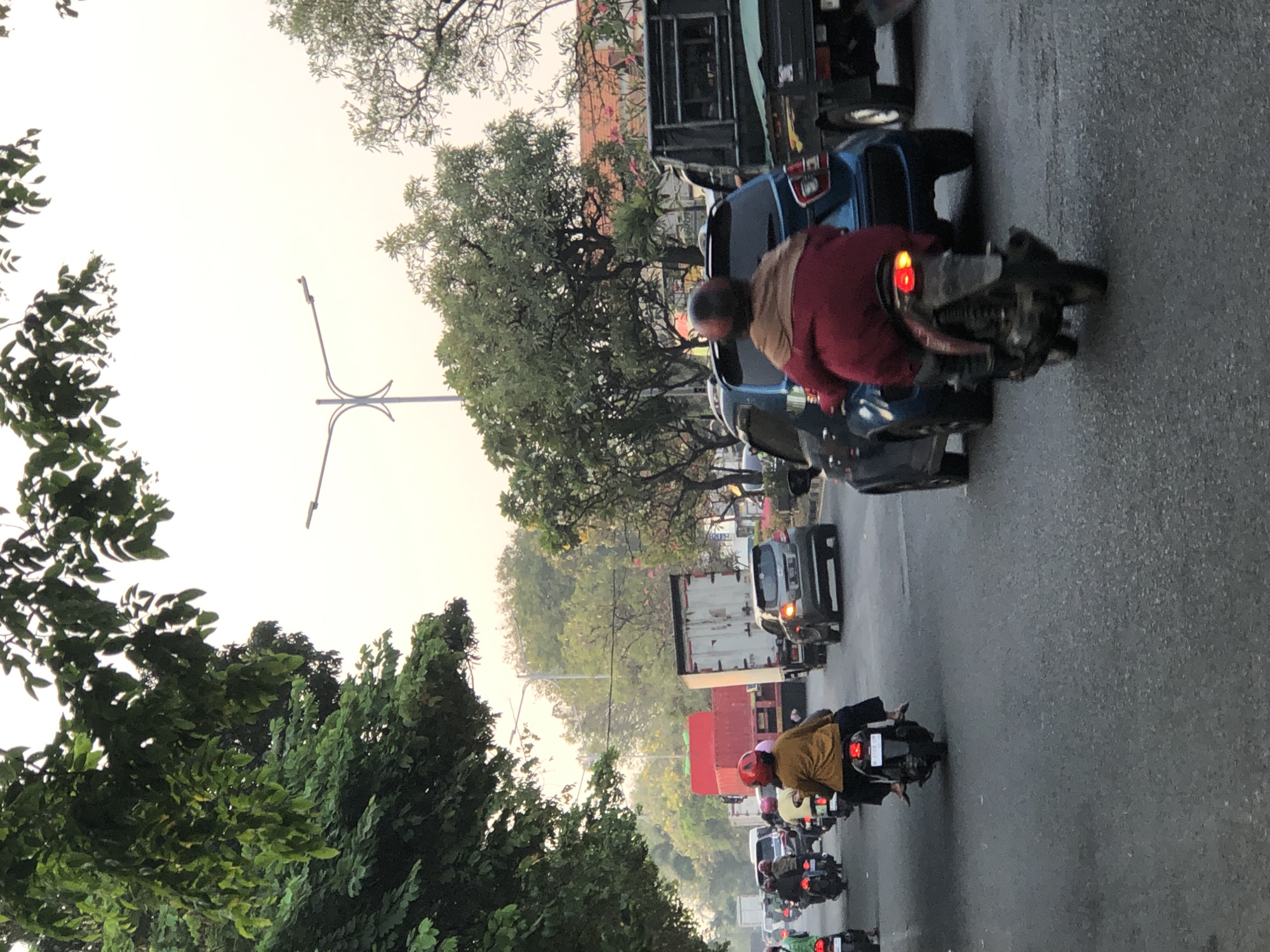 In Sumorame, Indonesia, a traffic jam occurs on a paved road with vehicles including motorcycles and cars. In the foreground, a person on a dark-colored motorcycle wears a red jacket and a brown scarf, with a red taillight illuminated. To their left, another person on a light-colored motorcycle wears a yellow top and dark pants. Behind them, a blue car is visible. Further down the road, a grey car is seen in traffic. Trucks, including a white one with a red cargo container, are also part of the congestion. Trees line the sides of the road, and a street light is visible in the distance. The sky is bright and overcast, suggesting it might be late afternoon or early morning.