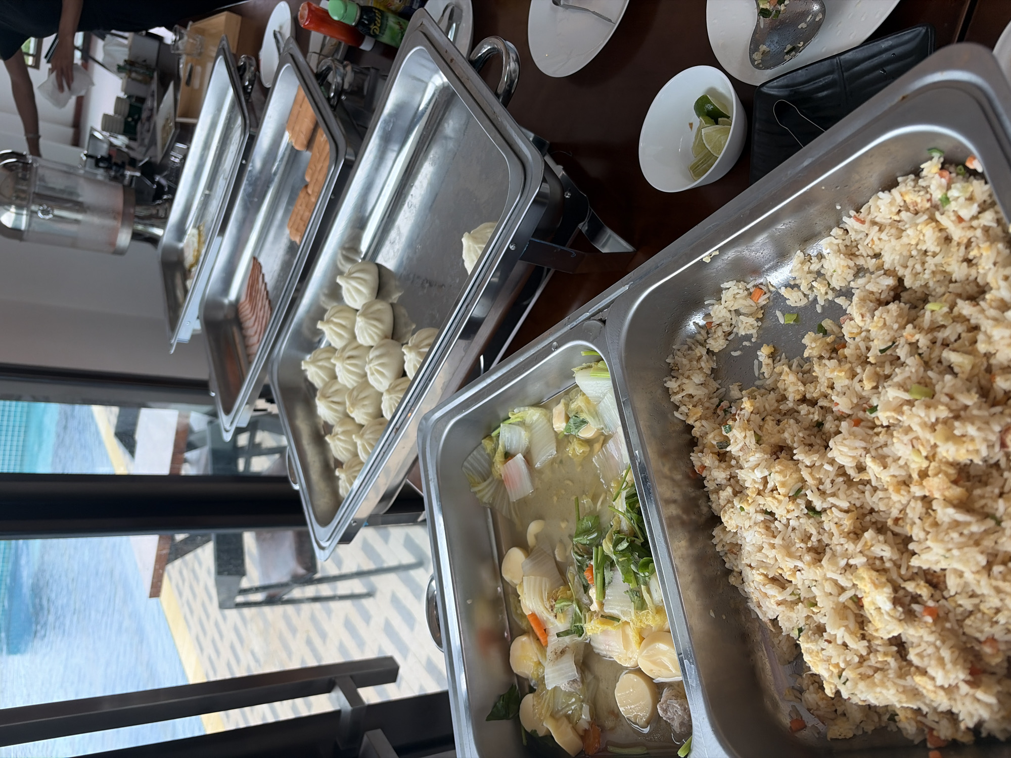 The image shows a buffet spread in what appears to be a resort or restaurant setting, possibly in Ban Kao Dua, Thailand, overlooking a swimming pool. In the foreground, a large stainless steel chafing dish contains fried rice with visible bits of egg and vegetables. To the left of the fried rice, another chafing dish holds a clear broth with pieces of tofu, cabbage, carrots, and what looks like ground meat.
Further back, several other chafing dishes are arranged on a table. One contains a row of steamed buns or dumplings. Another holds slices of bacon, and a third has hot dogs. In the background, a person in a dark shirt is visible near a beverage dispenser, holding a white cloth. The environment suggests a meal service, with plates and cutlery visible on the table. The outdoor pool and seating area visible through large windows indicate a location with a view, likely a resort or hotel.