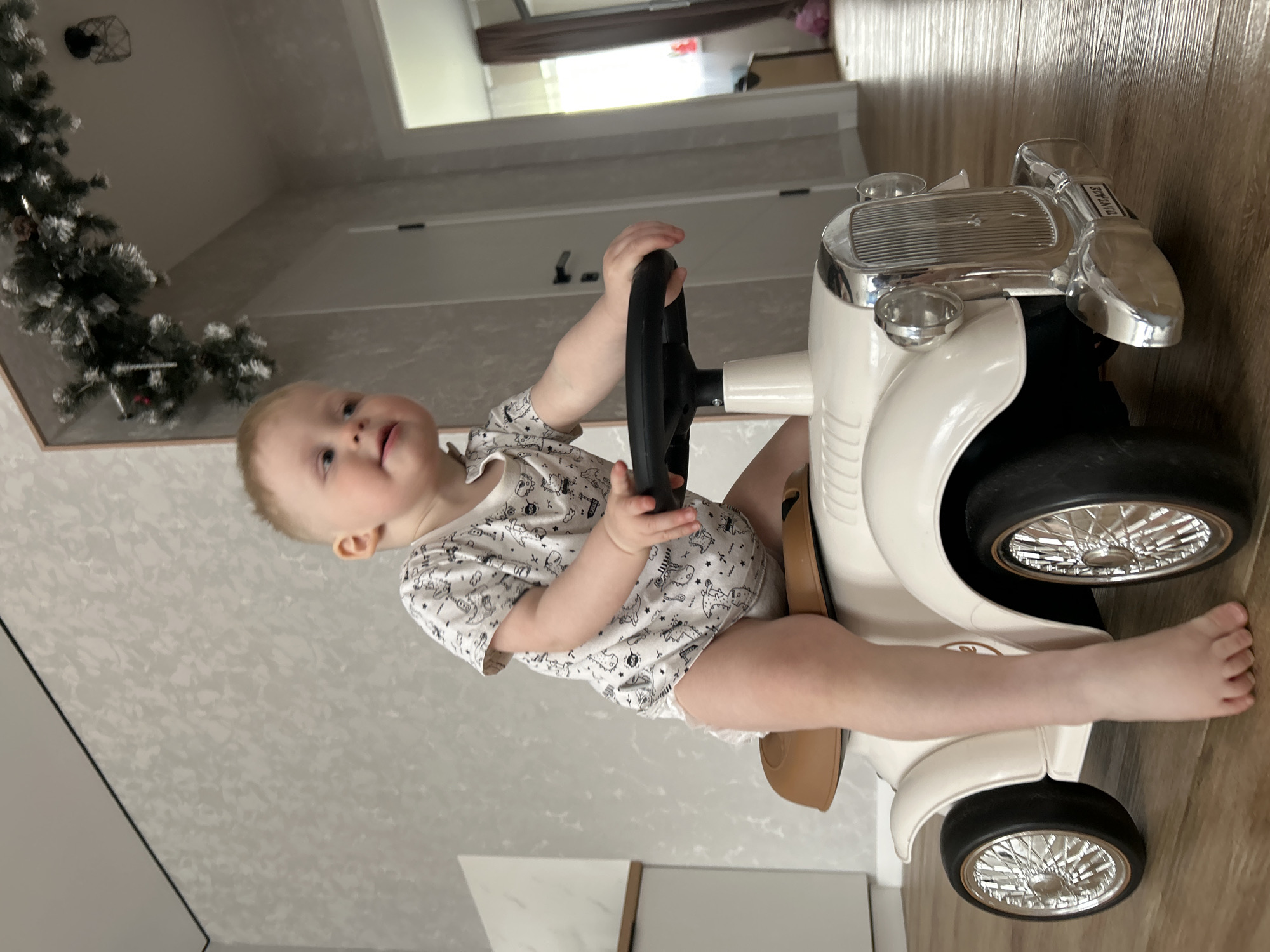A baby is sitting on a white toy car, holding the steering wheel. The car is on a wooden floor, and there is a decorated Christmas tree in the background. The scene appears to be indoors, possibly in a home. It is not possible to determine the location in the city of Chaikovsky, Russia from the given image.