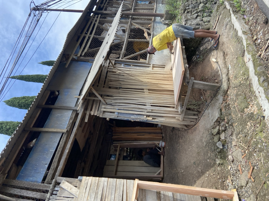 The image shows a man sanding a wooden door frame outside a woodshop in Rancabali, Indonesia, located at -7.123 latitude and 107.433 longitude. The woodshop appears to be a small, ramshackle building made of wood and corrugated metal. There are stacks of wooden planks and doors leaning against the walls. The man is wearing a yellow shirt and blue shorts. He is standing at a work table outside the building, using a power sander to smooth the surface of the door frame. The ground is dirt and gravel. There is a small, overgrown area to the left of the building, with a wooden fence surrounding it. There is a sidewalk in front of the building, with a curb on the right. The sky is blue and clear. There are power lines running overhead. There are a few trees in the background. The building is next to a steep hillside with a stone wall. The man is focused on his work. The scene is typical of a small town in Indonesia, with a mix of modern and traditional architecture. The man is in the process of creating a product, and the scene conveys a sense of craftsmanship and community.