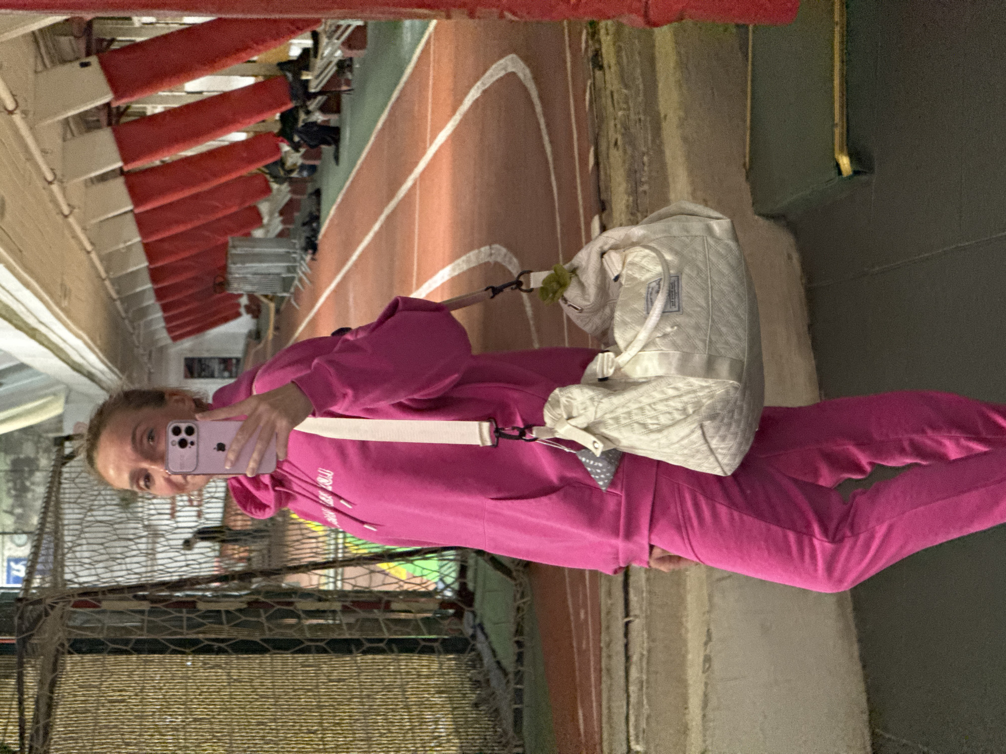 A young woman is taking a selfie with a pink mobile phone. She is wearing a pink hoodie and sweatpants, and has a white quilted purse slung over her shoulder. She is standing next to a handball goal on a sports field, specifically a running track inside a building. The location is identified as Murino, Russia. Behind the woman is stadium-style seating.