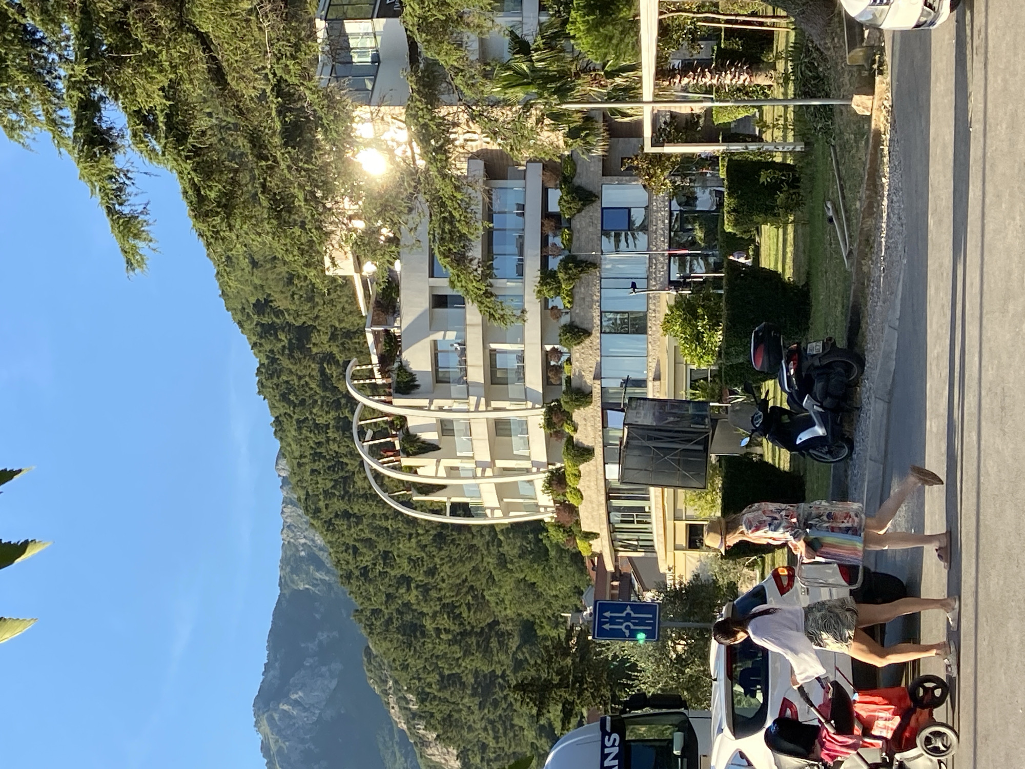 The image depicts a street scene in Budva, Montenegro, likely near the waterfront, characterized by a modern, multi-story building with arched balconies and extensive glass windows, reflecting the bright sunlight. The building is nestled against a backdrop of lush green mountains and trees, indicating a scenic coastal or mountainous region. In the foreground, two women are walking; one is pushing a stroller with a red bag, and the other is carrying a bag with a rainbow-colored pattern. They are dressed casually in summer attire. A white SUV is parked to their left, and a truck's cab is visible on the far left edge of the frame. A scooter is parked on the grass to the right of the women. A traffic signal showing a green light is positioned on a pole. The overall atmosphere suggests a warm, sunny day in a tourist-oriented area.