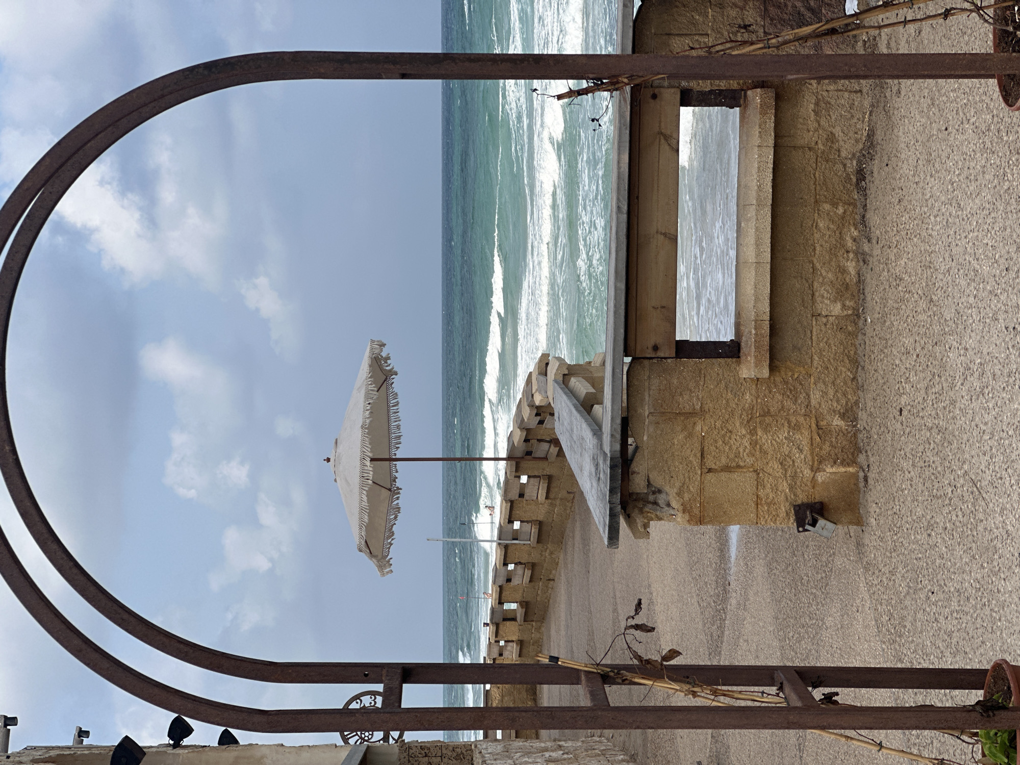This is a medium shot of a seascape, framed by a rusty metal archway. The archway is on the left side of the frame and extends upwards. Through the archway, a stone wall with wooden benches is visible, leading to the sea. The sea is a vibrant turquoise with white-capped waves rolling towards the shore. The sky is a mix of blue and white clouds. A large, white umbrella with fringes is set up on the right side of the wall, near the sea. There are a few people in the water, swimming or surfing. The foreground shows a paved walkway. The image appears to be taken in Netanya, Israel, based on its coastal scenery and architectural style.
