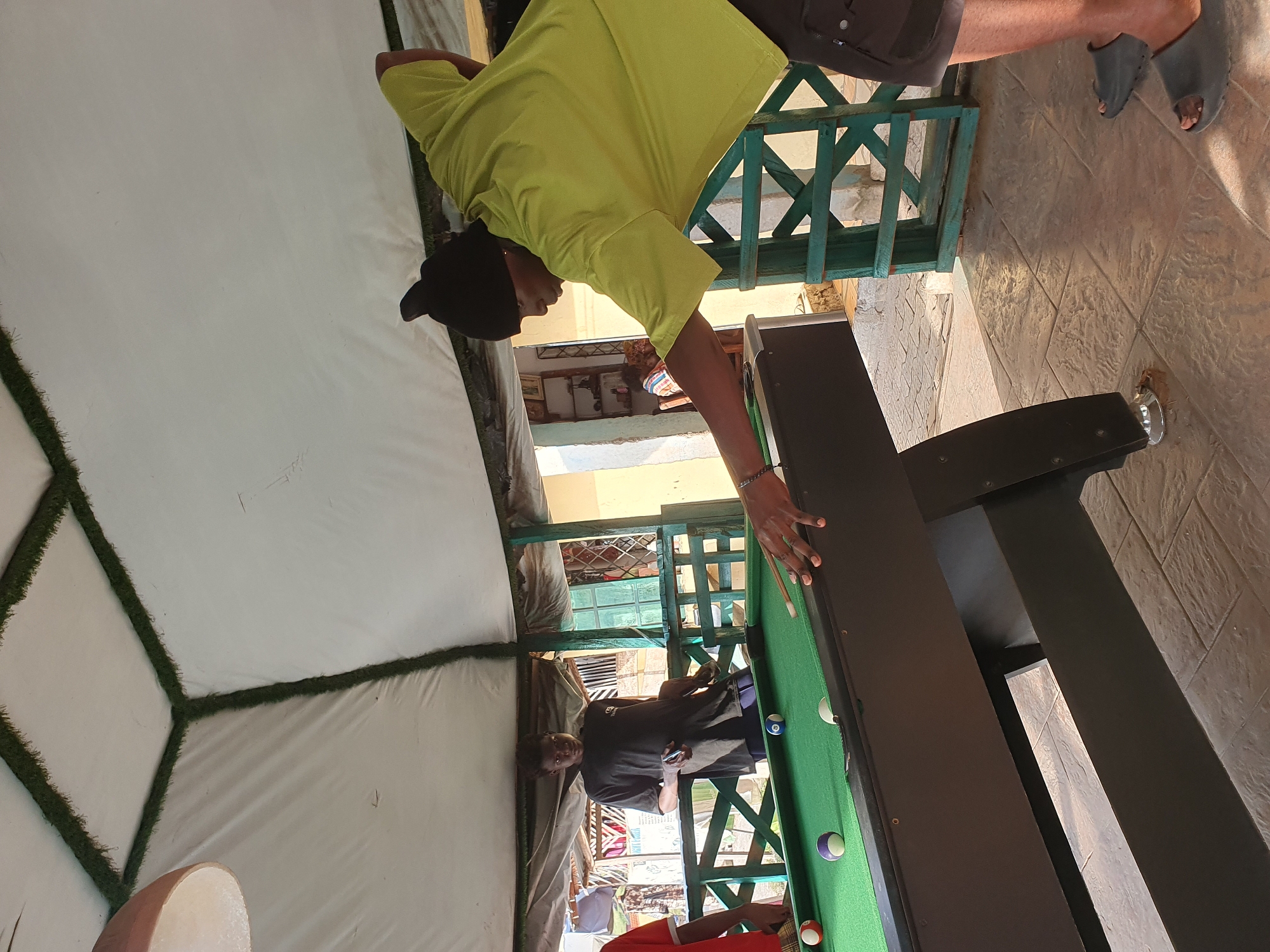 Two individuals are playing a game of pool at an outdoor establishment in Lokoja, Nigeria. The person on the right, wearing a bright yellow t-shirt and a black beanie, is in the process of taking a shot with a cue stick. The person on the left, dressed in a black t-shirt, is standing and observing the game. The pool table is green, with several balls visible on its surface. The scene appears to be under a canopy, with a partially visible light fixture and decorative green trim along the edge of the canopy. The ground is paved with patterned tiles. In the background, there are structures with wooden and metal elements, and some vegetation.