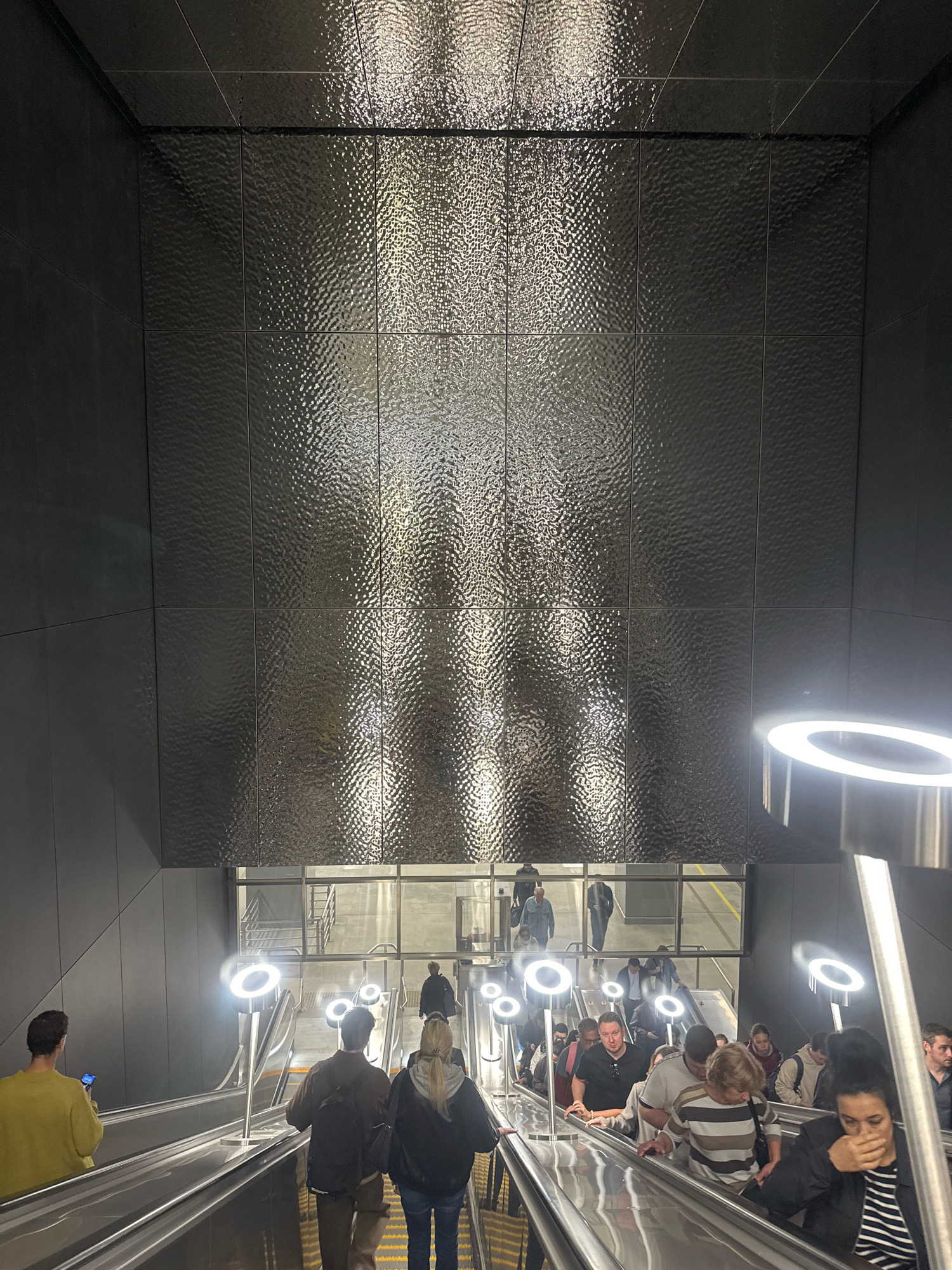 The image depicts an interior scene of a Moscow Metro station, likely a modern station with escalators and minimalist design. The focal point is a large, textured wall clad in dark, reflective tiles that create a rippling, almost liquid effect. Light reflects off these tiles in vertical patterns. Several escalators are visible, carrying people up and down. The escalators are flanked by contemporary lighting fixtures that emit a cool, white glow in a circular pattern. People are casually dressed and appear to be commuters or travelers. They are mostly walking on the escalators, some looking at their phones, others talking. The environment is clean and well-lit, suggesting a public transportation hub. The overall impression is one of efficiency and modern urban life.