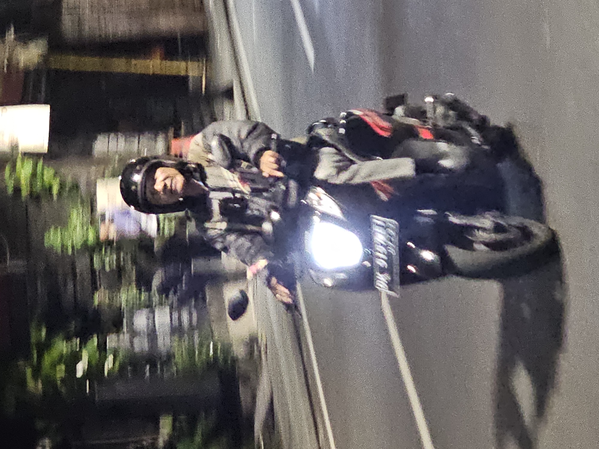 A person wearing a helmet and jacket rides a black motorcycle at night on a street in Denpasar, Indonesia. The motorcycle's headlight is on, and its license plate is visible. The background includes buildings and trees. The road appears to be asphalt.