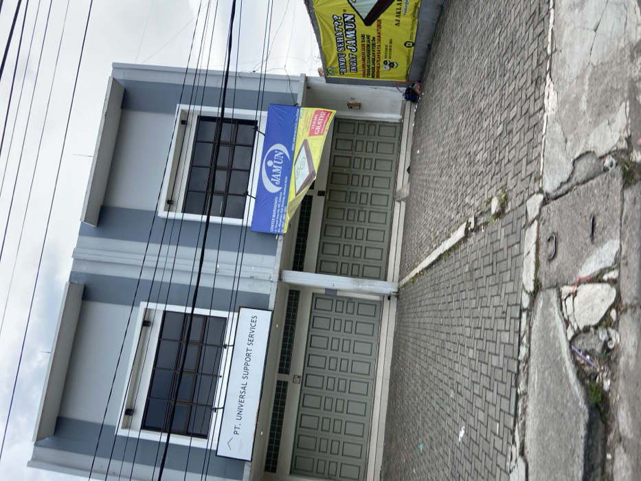 The image shows a street-level view of two commercial storefronts in Margahayu, Indonesia. The buildings are both constructed of concrete, with gray walls and gray tiled roofs. The storefronts have large, closed, green-painted wooden doors. The storefronts are located on a sloped paved driveway. The storefronts each have a sign above their doors, the first one reads "PT. UNIVERSAL SUPPORT SERVICES," and the second sign reads "CENTER MARGAHAYU JAM UN". The second storefront also has a larger sign, mostly yellow, that reads "RINDU SEHAT?? INGT JAMUN." The signs are visible against a bright sky with many power lines. A few plants and a small amount of trash are visible in the foreground.