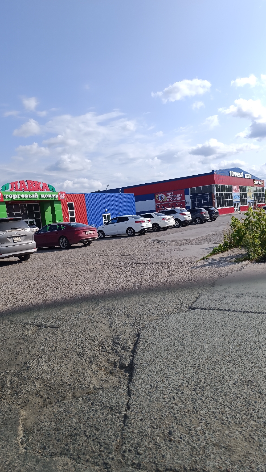 The image shows a commercial building with signage in Russian, located in the city of Chaikovsky, Russia. The building appears to be a shopping center, with "ТОРГОВЫЙ ЦЕНТР" (Shopping Center) visible on the facade. There are several cars parked in the lot in front of the building. The sky is partly cloudy, and there is some vegetation on the right side of the image. The foreground shows a rough asphalt surface with visible cracks. The overall scene depicts a typical urban commercial area on a sunny day.