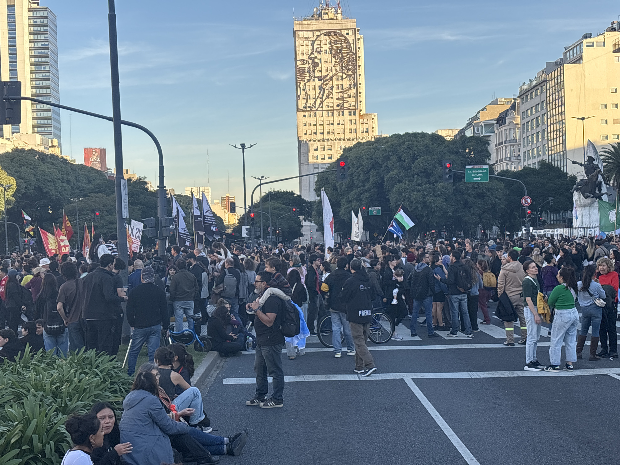 The image shows a large crowd of people gathered on a wide street, likely for a protest or demonstration. The location appears to be in Buenos Aires, Argentina, based on the distinctive architectural style of the buildings in the background. Prominently visible is a high-rise building with a large mural of a woman's face. The crowd is dense and diverse, with people of varying ages and appearances. Many individuals are carrying flags, signs, or banners. Some people are standing, while others are sitting on the ground. The street is marked with crosswalks, and traffic lights are visible, indicating the location is an intersection. The atmosphere seems active. The weather appears clear and bright, with a blue sky.