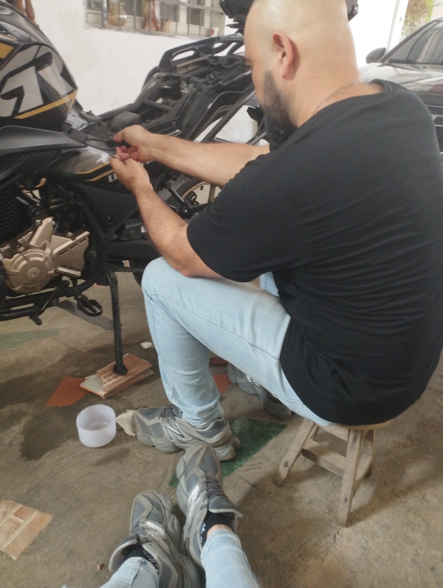 In a casual setting, likely a garage or workshop in Tinaquillo, Venezuela, a bald man with a beard, wearing a black t-shirt and light blue jeans, is actively engaged in working on a motorcycle. He is seated on a small wooden stool, leaning forward, and using tools to perform maintenance or repair on the black motorcycle, specifically on its upper components near the fuel tank and handlebars. A silver chain is visible around his neck.
The motorcycle, which features black, gold, and white accents with graphics resembling "T R" and "LI" or "LU", is propped on its kickstand. The kickstand is resting on a stack of reddish-brown bricks or tiles for added stability or elevation on the concrete floor. The floor itself appears worn and somewhat uneven.
In the foreground, the legs and feet of another individual (presumably the photographer) are visible, clad in light blue jeans and grey sneakers, propped up as if resting or observing the work. A white plastic cup sits on the concrete floor near their feet.
The background shows a white wall with a window covered by a metal grate. A dark-colored car is partially visible behind the motorcycle, suggesting it's parked nearby. The lighting suggests it is daytime, possibly indoors or under a covered structure. The scene conveys a focused, hands-on repair activity.