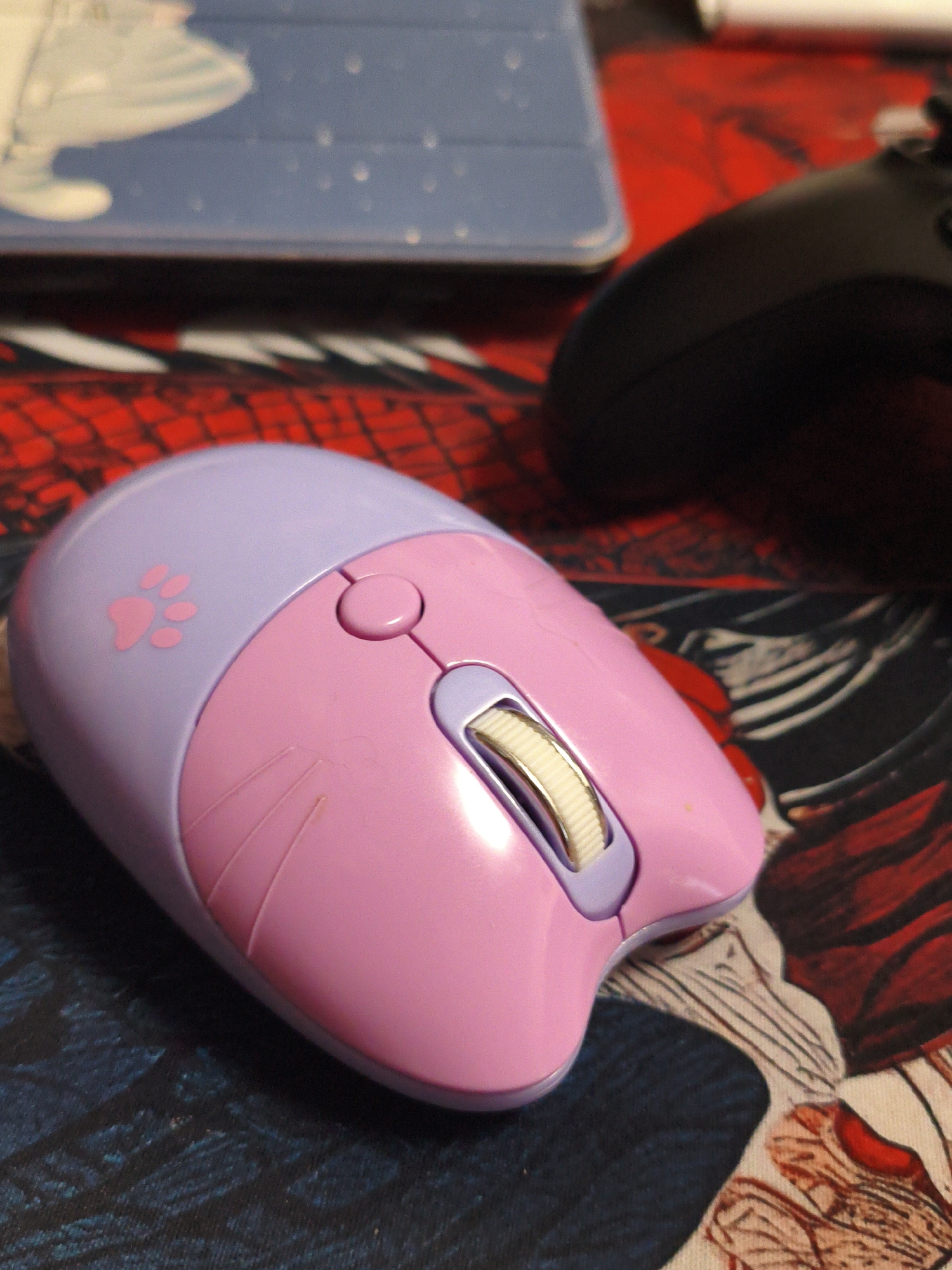 A pink and purple computer mouse with a paw print design is positioned on a patterned mousepad. In the background, a portion of a blue tablet and a black gaming controller are visible. The scene appears to be indoors, likely on a desk. There are no people present in the image. The image does not provide any information about the city of Vladivostok, Russia.
