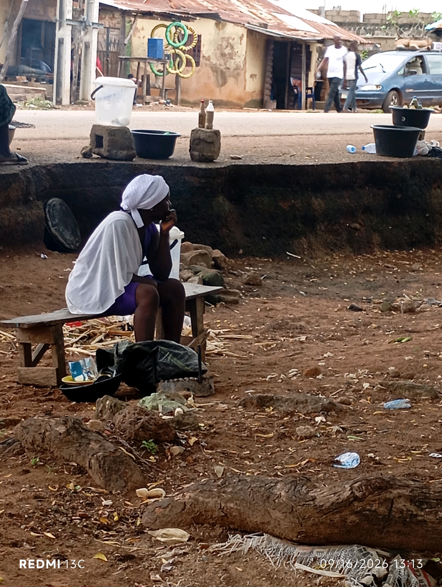 In Shendam, Nigeria, a woman wearing a white headscarf and a purple dress sits on a wooden bench. She is looking to her right and appears to be eating or resting her hand on her chin. In front of her, on the ground, are various items including a black bowl with food, a black bag, and other debris. To her left, a black bowl is placed on a makeshift stand. In the background, a road is visible with a few people walking and a blue car parked. The environment appears to be a roadside market or informal settlement, characterized by simple structures and a dusty, unpaved ground. The overall scene depicts a moment of everyday life in this Nigerian city.