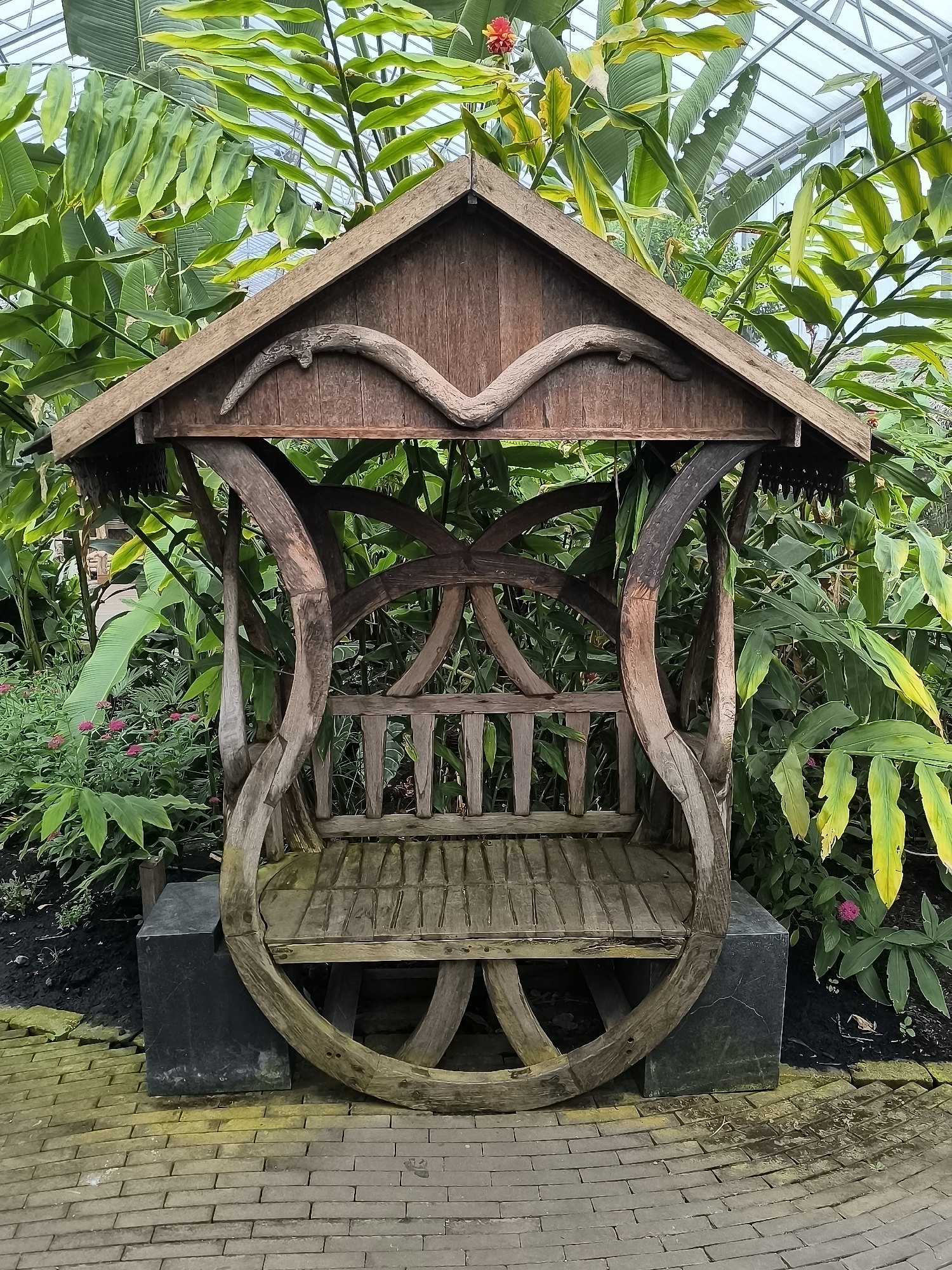 A full-shot photograph depicts a wooden structure resembling a covered bench in a lush greenhouse setting. The structure features a pitched roof with decorative wooden carvings and a curved support system forming the seat's base. The bench seat is made of slats. It rests on two square dark gray stone blocks on a brick-paved floor. Behind the bench, dense foliage creates a green backdrop, suggesting a tropical or subtropical environment. The roof of the greenhouse is visible at the top of the frame. The location is Waarland, Netherlands. There are no people visible in the image.