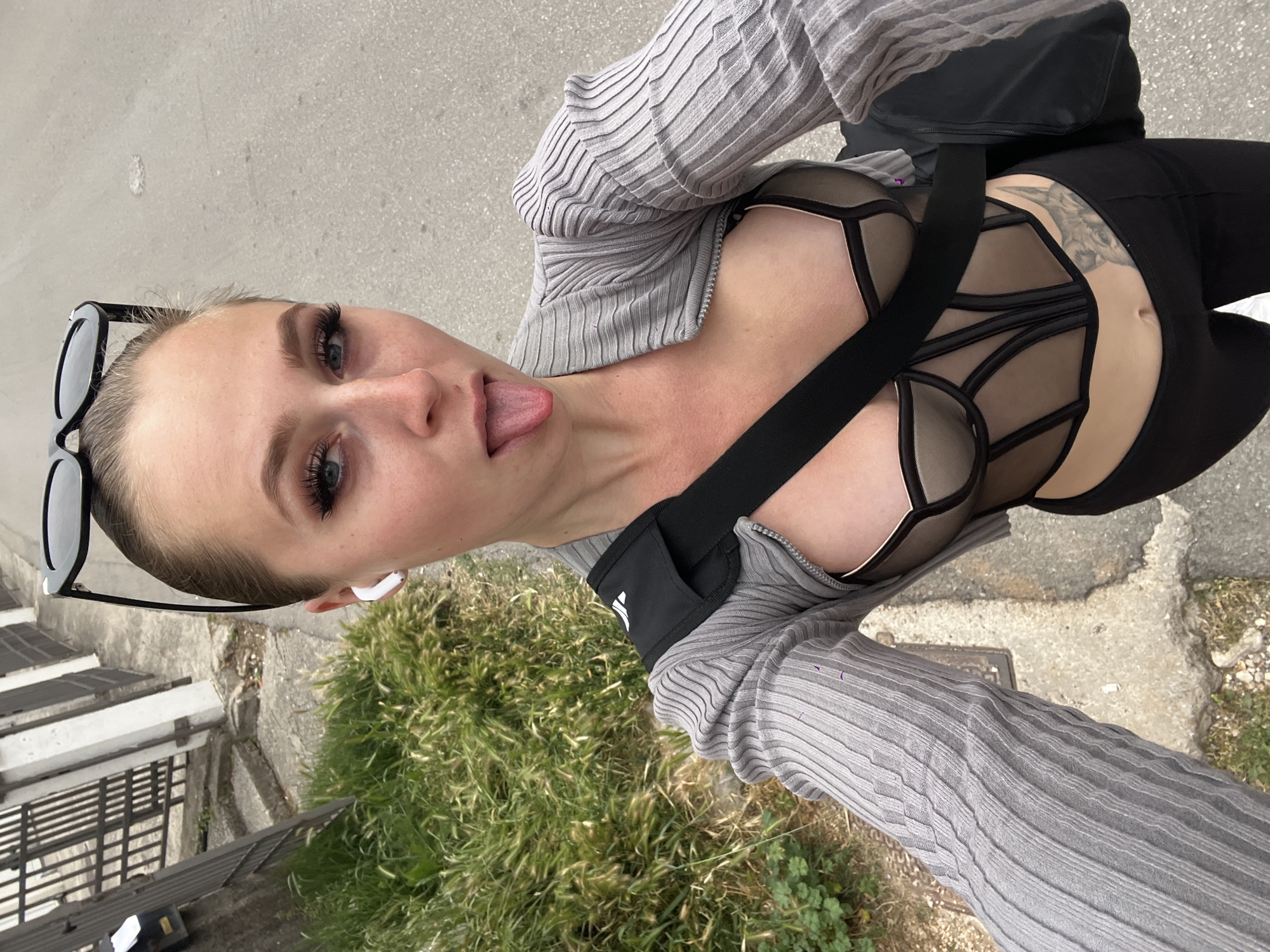 A woman poses for a selfie outdoors. She sticks her tongue out and wears black leggings, a black bra-like top with straps and geometrical designs, and a grey ribbed long-sleeved shirt. A black shoulder bag's strap crosses her body. Sunglasses are perched on top of her head, and she has an earbud in her right ear. In the background, there is green vegetation and a concrete sidewalk. The setting may be in Boreti, Montenegro, given the provided context.