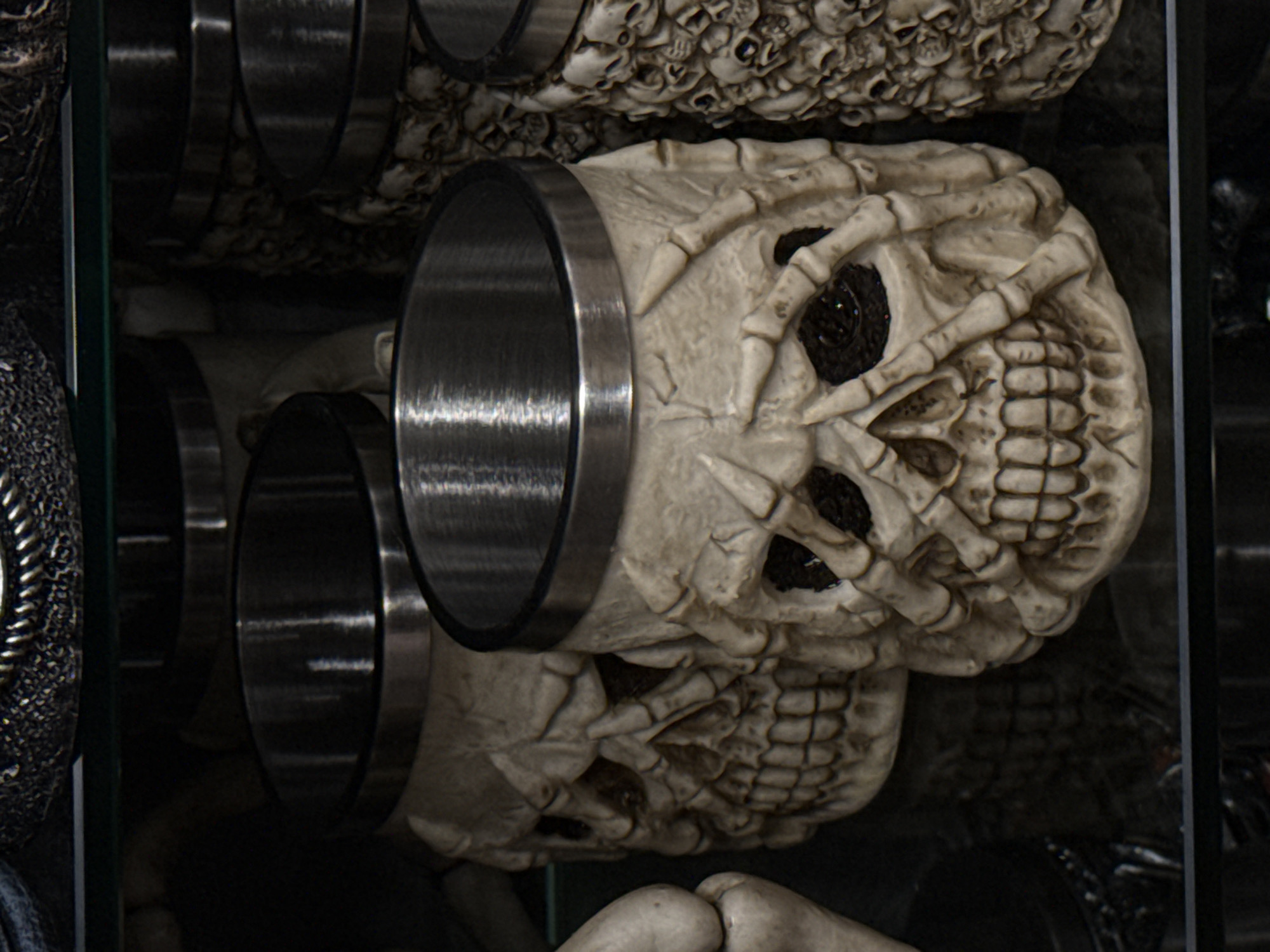 This image depicts a collection of skull-themed drinking vessels with metallic rims, displayed in Primorsko, Bulgaria. The central vessel, in sharp focus, is an off-white skull-shaped tankard featuring articulated skeletal hands gripping its facial structure, particularly around the dark-recessed orbital sockets. The mandible displays a full set of teeth, and a brushed metal band forms the rim of the opening. To the immediate left, a portion of a similarly textured, bone-like object is visible. To the right and slightly behind the central item, another tankard is present, its surface intricately patterned with multiple small, relief-sculpted skulls. A third, partially obscured skull tankard is discernible behind the central item. All items rest on a dark, highly reflective surface, which shows discernible reflections of the metallic rims and lower portions of the vessels. A thin, horizontal green line, indicative of a glass shelf or barrier, spans across the upper background. The overall illumination is subdued, highlighting surface textures and metallic reflectivity against a dark background.