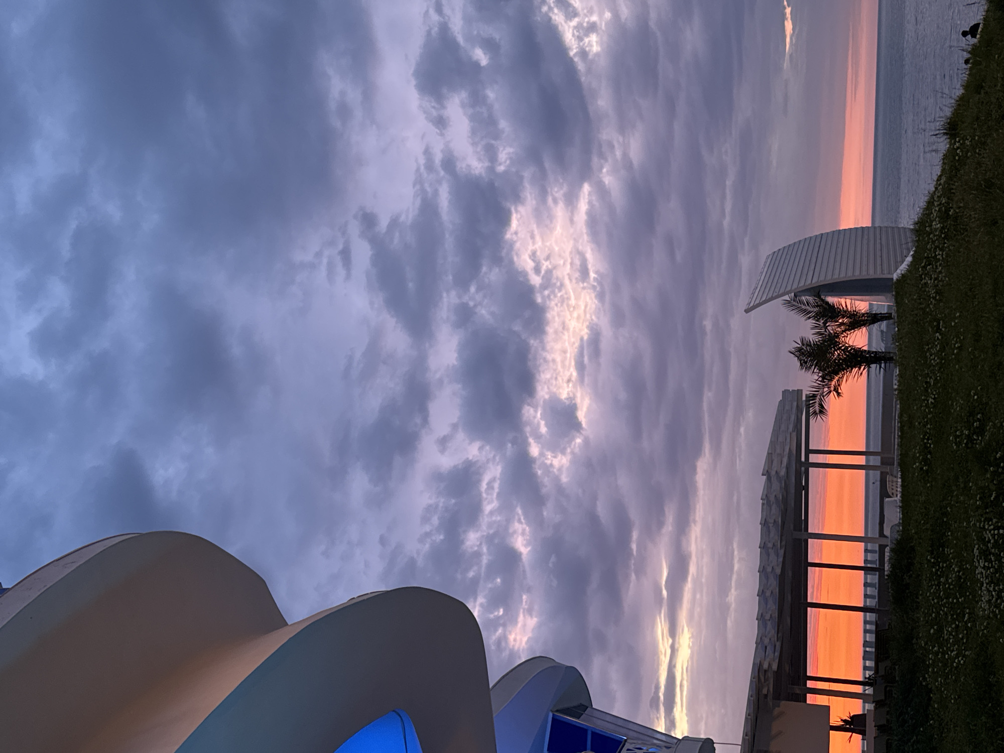 The image shows a coastal scene at sunset. The sky is filled with purple and grey clouds, with hints of orange near the horizon. The sea is visible as a thin strip in the distance.
In the foreground, there are architectural structures on either side. On the bottom left is part of a modern building with curved white walls and blue lighting. On the right is a beachside structure with a corrugated roof and supporting beams, with an orange glow illuminating the space beneath, presumably from the setting sun. A palm tree is silhouetted against the sunset behind this structure. The location is Unknown, Unknown.