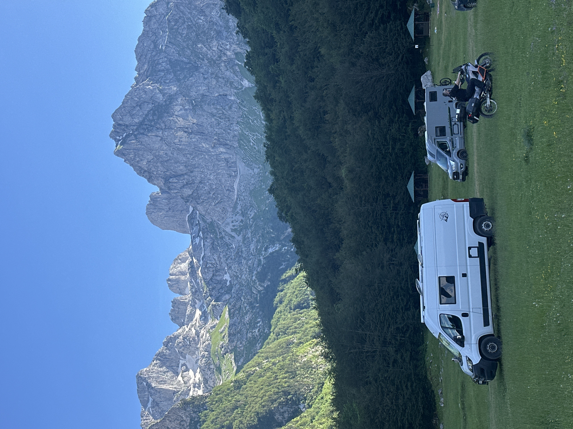 In the city of Dolja, Montenegro, a scenic camping area is depicted under a clear blue sky. The background is dominated by majestic, rugged mountains with patches of snow, characteristic of the Prokletije range. A dense, green forest covers the lower slopes of the mountains and extends to the mid-ground of the scene. In the foreground, a wide, grassy field is dotted with several vehicles and small wooden structures.
A white camper van is parked on the left side of the field. To its right, a grey camper van is situated. A person is riding a motorcycle in the middle of the field, heading away from the viewer. There are at least three small, green, pagoda-style shelters scattered across the field. The overall environment suggests an outdoor recreational space, likely a campground or a scenic viewpoint, where people are engaged in leisure activities.