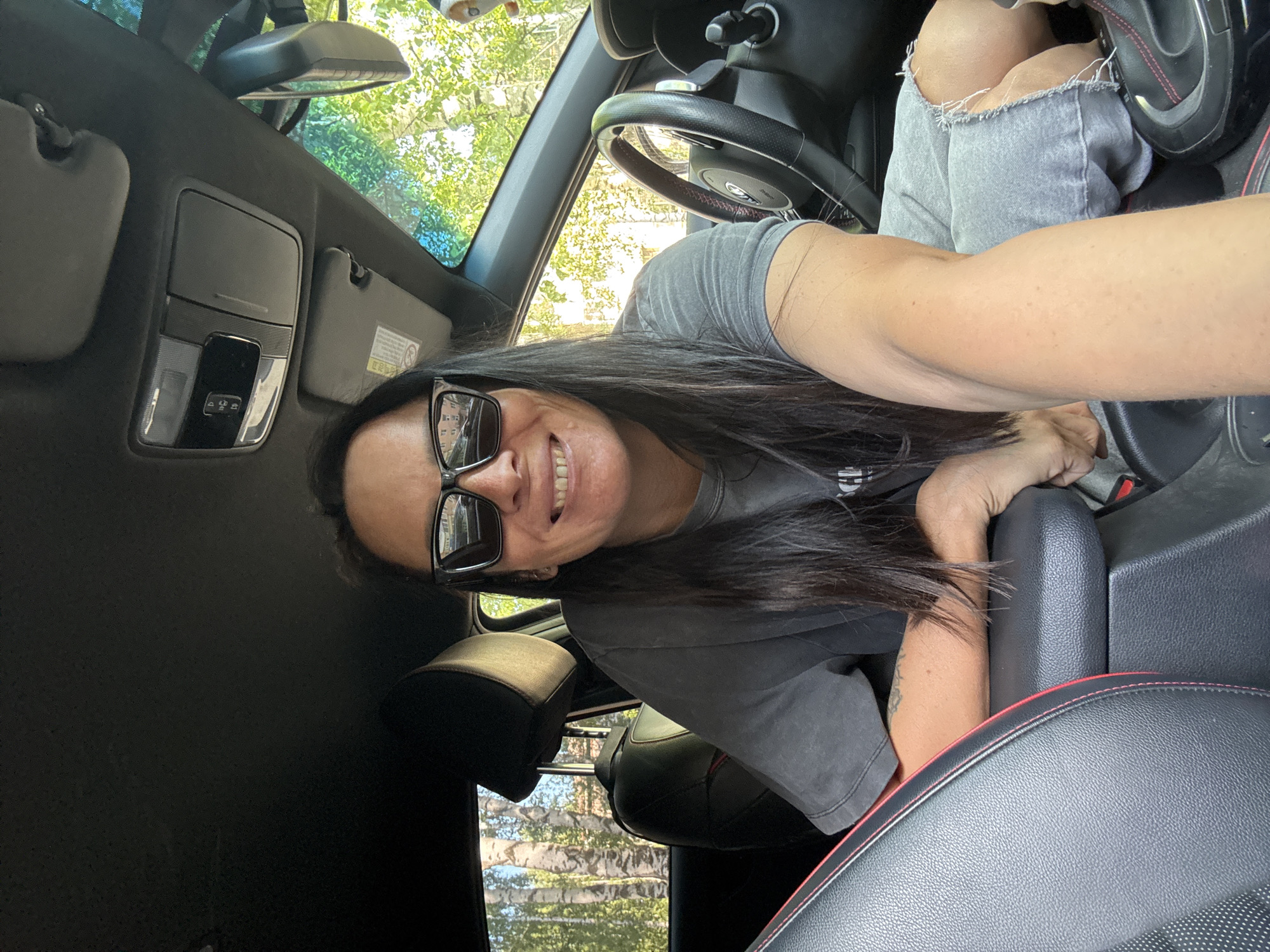 A person wearing sunglasses and a gray t-shirt is in the driver's seat of a car, with the steering wheel and dashboard visible. The interior of the car features black leather seats with red stitching. Outside the car, trees can be seen through the windows. The location of the scene is unknown and not deducible from the image.
