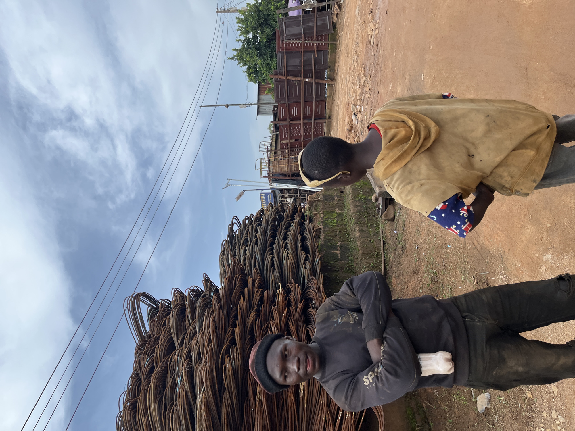 This media file captures a scene outdoors, likely in Gura Top, Nigeria. Two young individuals are the primary subjects. The person in the foreground wears a dark sweatshirt with visible text, "SPA" and "SEM" partially obscured. They also wear a dark beanie with a red band and black pants. Their arms are crossed, and they are looking towards the camera with a neutral expression. A large pile of rebar, commonly used in construction, is prominently displayed to the left of this individual, dominating a significant portion of the frame.
The second individual is seen from the side, wearing a light-colored, possibly tan or yellow, jacket over a red shirt. They have a patterned blue and white cloth wrapped around their head or neck. This person is looking away from the camera, towards the background.
The background reveals a dusty, unpaved ground and hints of a market or industrial area. Stacks of metal structures, possibly gates or fencing, are visible further back, along with some greenery. Overhead, power lines crisscross the sky, which is partly cloudy with white wispy clouds against a blue expanse. The lighting suggests it is daytime, but the exact time is not clearly discernible. The overall impression is of a working environment, possibly a junkyard or a construction material supplier.