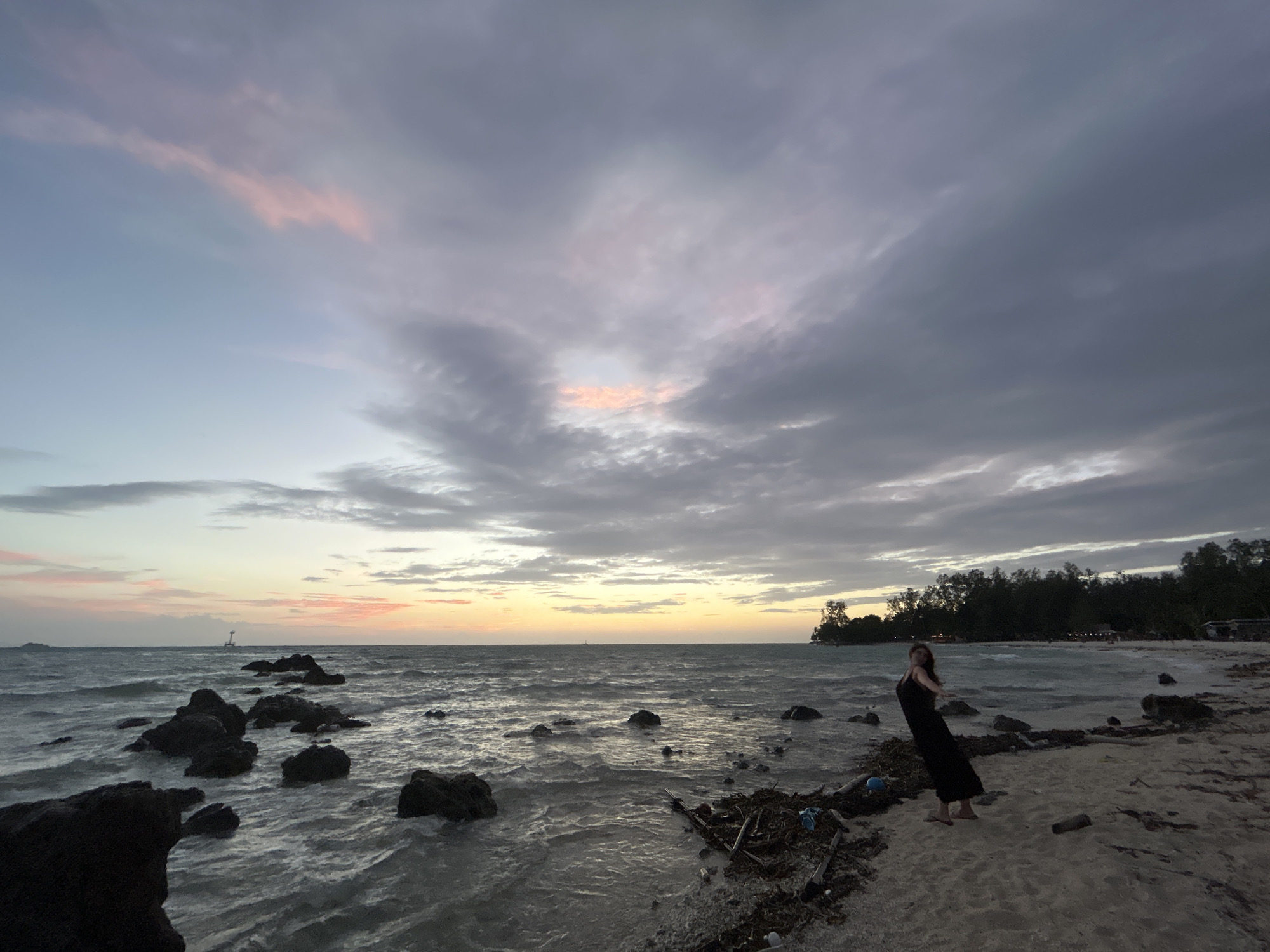 A lone individual wearing a long black dress stands on a sandy beach, her body leaning back with arms outstretched. The scene is a coastal environment in the Ko Pha-ngan District, Thailand, likely during twilight or dawn. The sky above is filled with dramatic, cloudy formations, interspersed with soft hues of pink and orange, indicating the sun is either rising or setting. The sea is a muted gray, with gentle waves lapping at the shore. Scattered rocks are visible both in the water and on the beach, along with debris such as driftwood and plastic. In the distance, a line of trees forms the backdrop, with faint lights suggesting human habitation. A small structure or beacon is visible on the horizon over the water. The overall mood is serene and slightly melancholic, with the natural beauty of the coastal landscape being the dominant element.