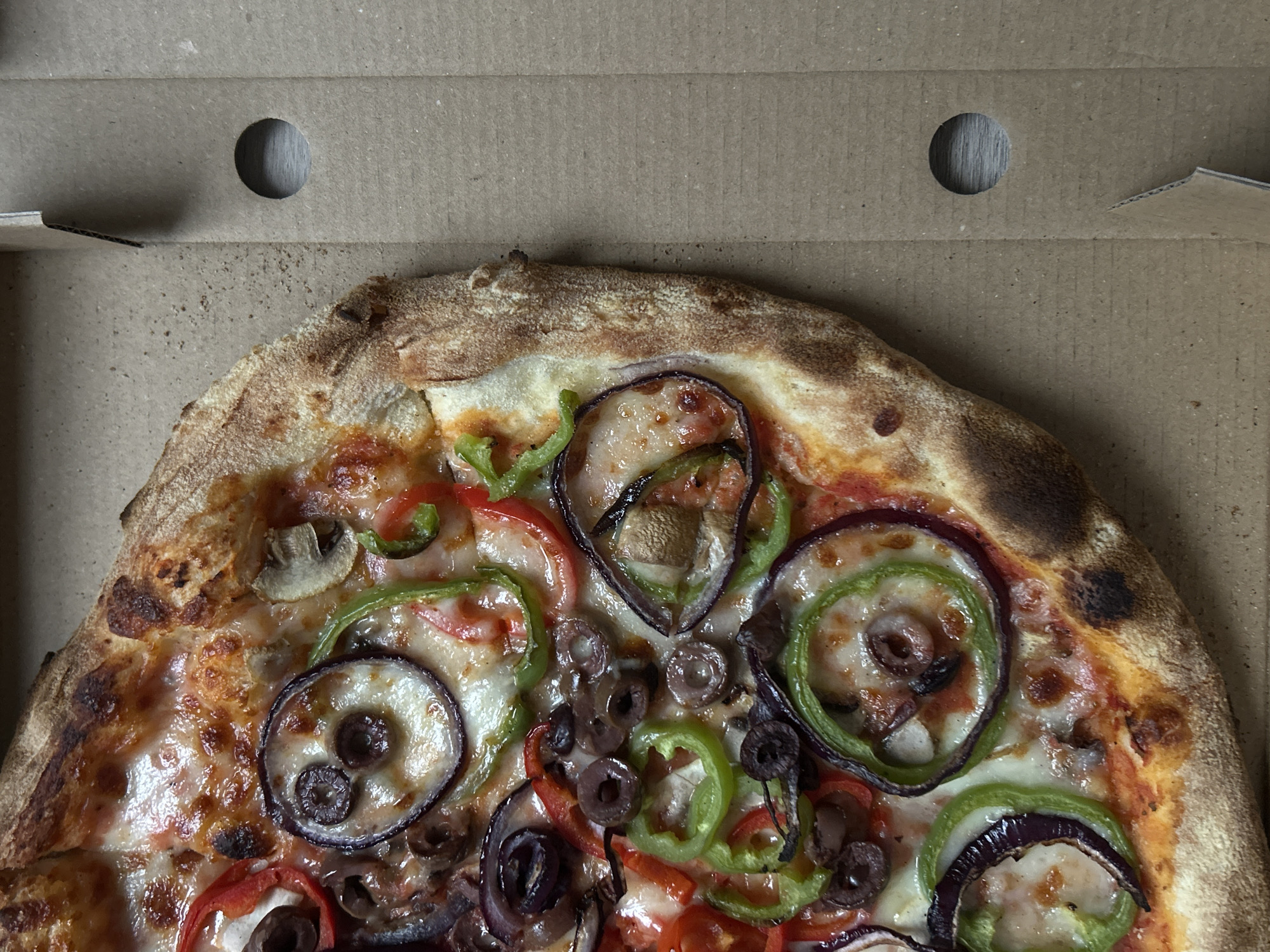 The image presents an overhead, close-up view of a vegetarian pizza inside an open, light brown corrugated cardboard delivery box. The pizza occupies the lower two-thirds of the frame, with the interior of the box lid visible at the top.
The pizza has a thick, well-baked crust with some darker, charred areas, suggesting a hot oven bake. Its surface is generously covered with melted, bubbly cheese, partially revealing a red tomato sauce base underneath. A variety of fresh toppings are distributed across the cheese. These include prominent concentric rings of sliced red onions and bright green bell peppers. Slices of red bell pepper, black olives (some whole, some sliced), and sliced mushrooms are also clearly visible, creating a colorful and appealing arrangement.
The pizza box itself is made of standard cardboard material. Two circular ventilation holes are visible in the upper section of the box's lid, designed to allow steam to escape. A folded cardboard flap is also seen on the left side of the box.
No people are visible in the image, and there is no discernible activity other than the presentation of the pizza in its box, ready for consumption. There is no visible text or branding on the pizza or the box. The scene depicts a freshly prepared pizza, with the context provided indicating a location in Sofia, Bulgaria.