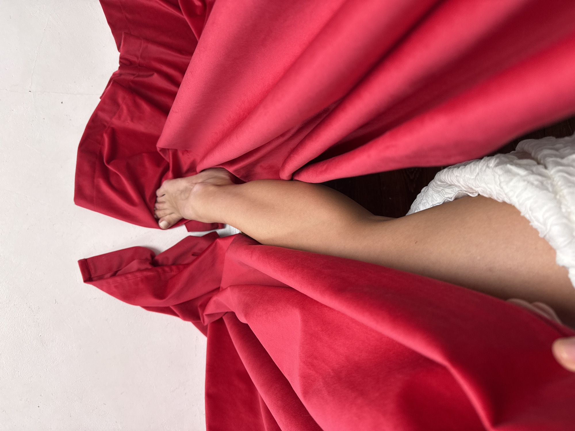 The image shows a person's leg and foot emerging from a draped red fabric. The person is wearing a white textured garment. The background includes a white wall and a dark wooden floor. The image does not contain information that allows me to determine a location in Chaikovsky, Russia.