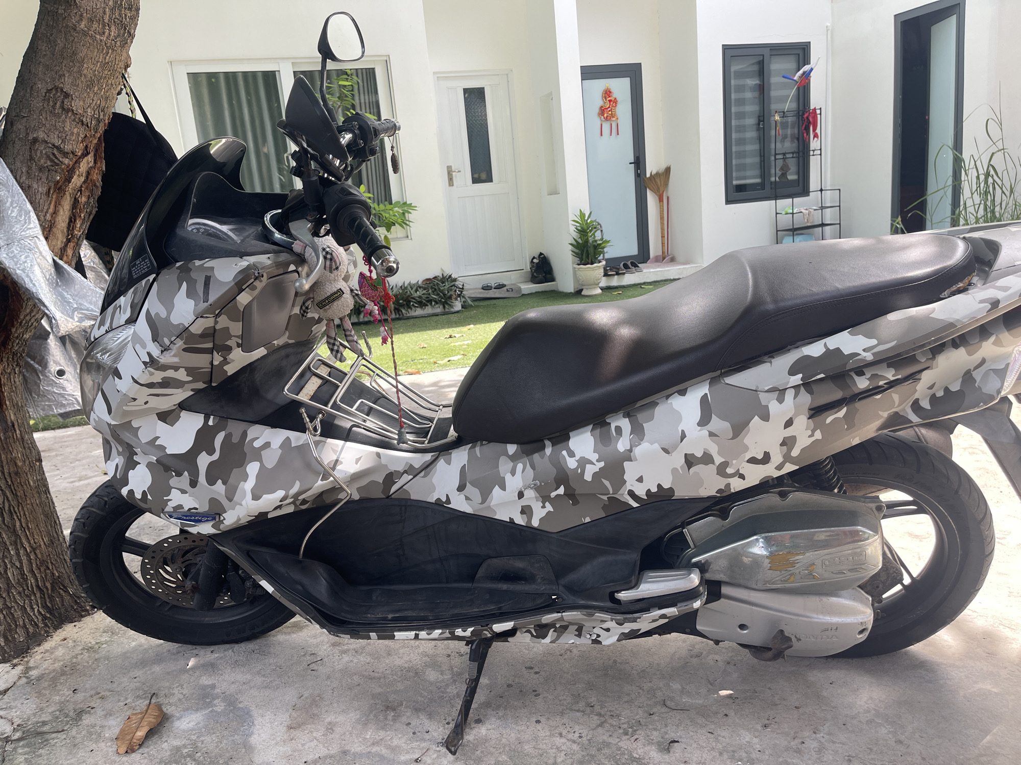 A camouflage-patterned scooter is parked on a concrete surface next to a tree trunk. The scooter has a black seat and a metal rack behind it. In the background, a white house with large windows and a grey door is visible. A green lawn with potted plants and a broom leans against the house. The scene appears to be a residential area in Phu Thuy Ward, Vietnam.