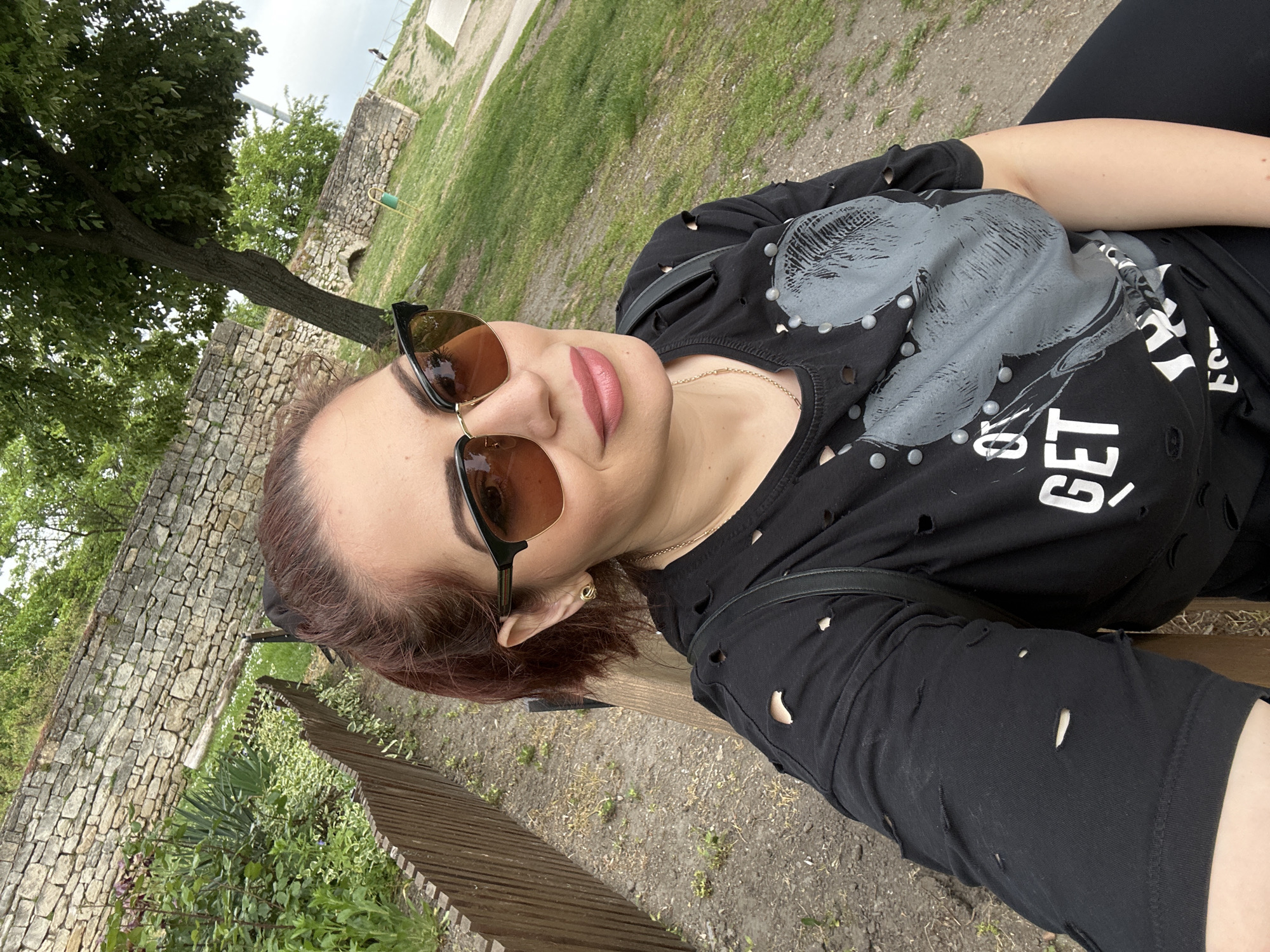 A woman is seated on a bench in an outdoor setting. She is wearing sunglasses and a black t-shirt with embellishments. The scene appears to be in a park or garden setting with a stone wall and trees visible in the background. The location is likely in Vidin, Bulgaria.