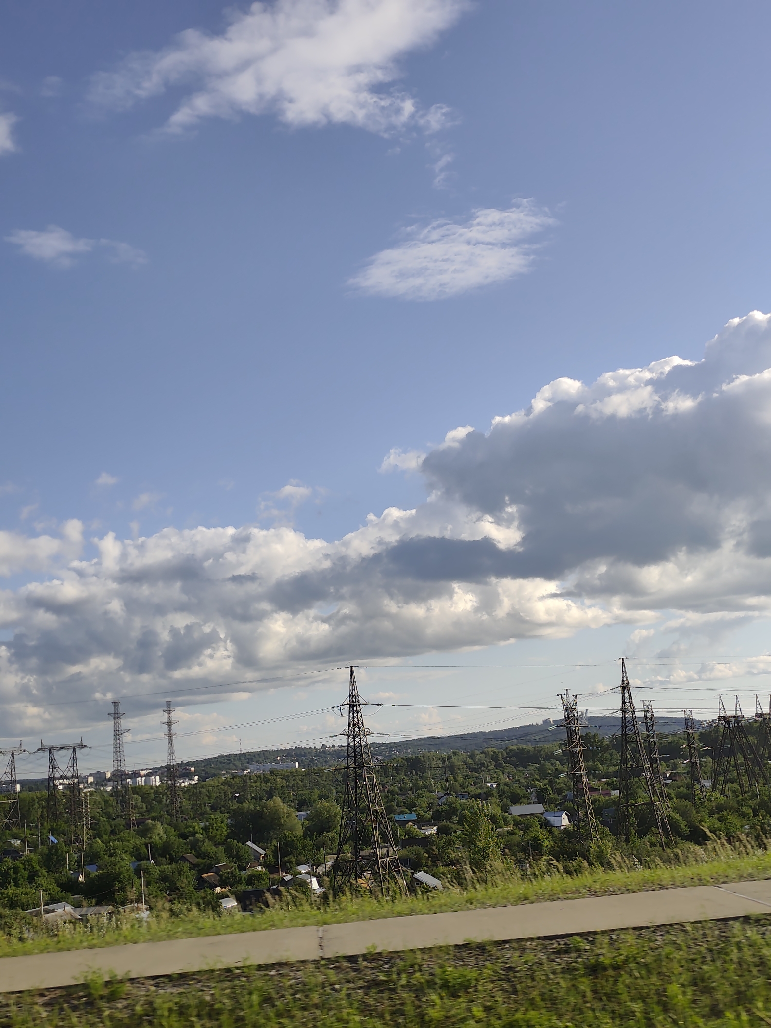 The image depicts a landscape with power transmission towers and a small settlement in the city of Chaikovsky, Russia. The sky is blue with scattered white and gray clouds. In the foreground, a strip of green grass and a paved path are visible. In the middle ground, numerous tall, dark metal power transmission towers stand amidst lush green trees and scattered houses, indicative of a suburban or rural area within the city. The settlement appears to be a collection of low-rise buildings, possibly individual homes or small cottages. The background shows a slightly hilly terrain with more trees and distant structures, suggesting the edge of the urban area. The lighting suggests it is daytime, likely afternoon, given the shadows and cloud formations. There are no people or animals visible in the image.