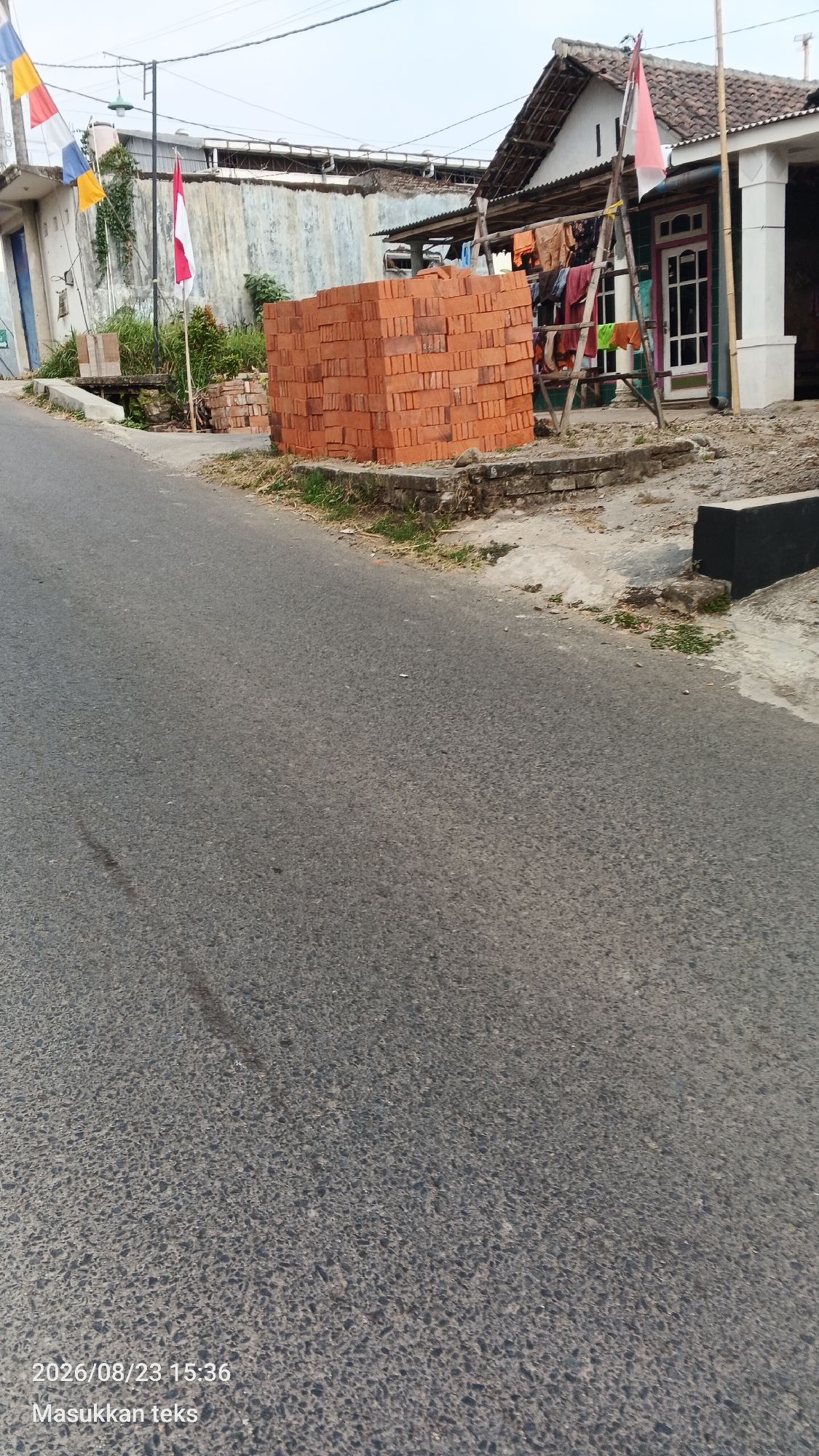 The image depicts a roadside scene in the city of Jeru, Indonesia. A stack of orange bricks is prominently placed on a concrete surface adjacent to an asphalt road. To the left, a white building with a blue doorway stands beside a canal or drainage ditch. In the background, a long, weathered concrete wall is visible, with an Indonesian flag flying from a pole in front of it. Further to the right, a house with a thatched roof and an open-sided structure displaying laundry on a clothesline is situated. The sky is overcast. The foreground consists of the asphalt road. A timestamp and text overlay in Indonesian, "2026/08/23 15:36 Masukkan teks," are present in the lower left corner of the image.
