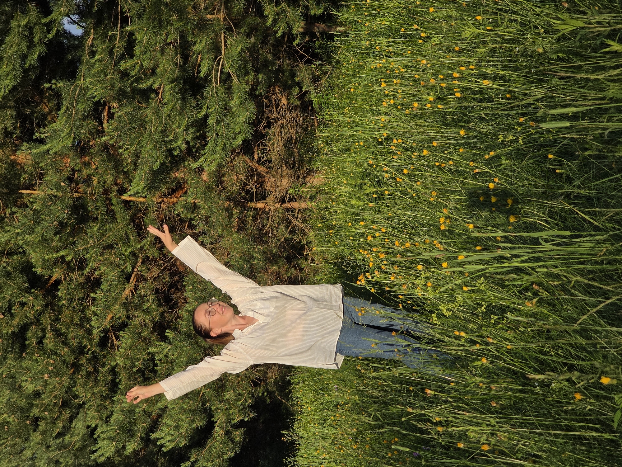 A young woman stands in a meadow filled with tall green grass and small yellow flowers, with her arms raised in a gesture of joy or celebration. She is wearing a loose white striped shirt and blue jeans, and round glasses. Behind her is a dense forest of pine trees. The sun is casting a warm glow on the scene, indicating it is either early morning or late afternoon. The specific location of Chaikovsky, Russia, is not determinable from the image itself, but the natural setting suggests an outdoor environment, possibly a park or a rural area.