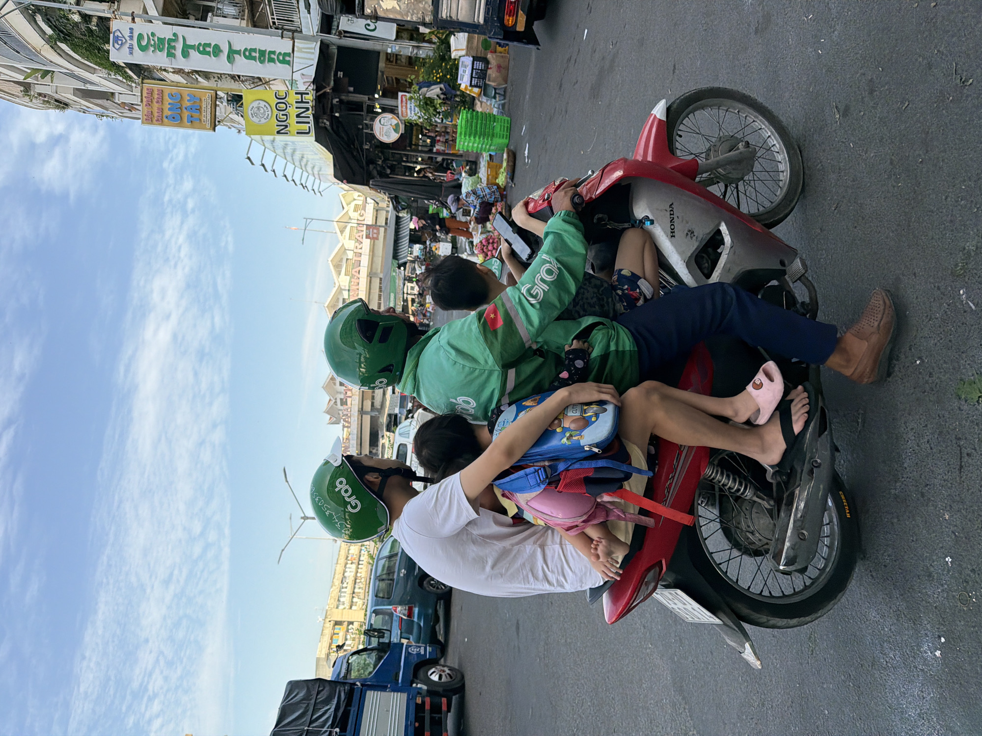 In Nha Trang, Vietnam, four individuals are riding on a single red and silver Honda motorcycle. The person in the front is wearing a green Grab helmet and a green jacket with the Grab logo. They are operating the motorcycle and looking forward. Behind them, a person in a white t-shirt is seated, also wearing a green Grab helmet. This person is holding a blue backpack with cartoon characters on it. Two children are seated between the two adults. The child in front of the person in the white t-shirt is wearing a pink shirt and pink flip-flops. The child behind them is wearing a patterned shorts. The motorcycle is on a paved road with other vehicles and buildings visible in the background. The sky is clear with some clouds. A blue truck is parked to the left of the motorcycle. Signs in Vietnamese are visible on the buildings.