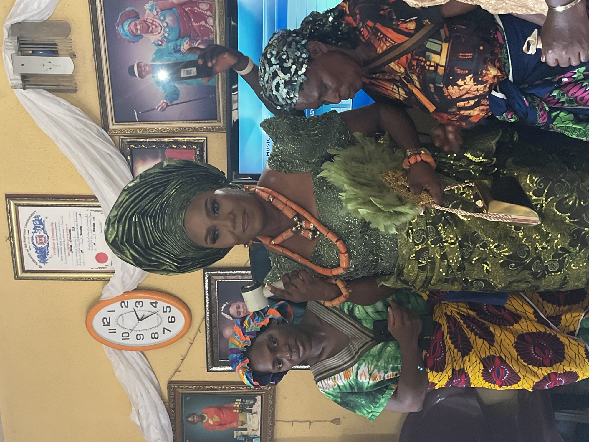 Here is a detailed description of the image:
**General Overview:**
The image captures a scene likely from a special occasion or event. It includes a woman dressed in elaborate traditional attire, possibly a bride or a celebrant, being attended to by two other women. The setting appears to be indoors, with walls adorned with pictures and decorations.
**People:**
1. **Woman in Green:** The focal point of the image is a woman wearing a green outfit with intricate beadwork and an elaborate headwrap of the same color. She is adorned with coral beads around her neck and wrist and holding a feathery fan.
2. **Woman Attending (Head Scarf):** An older woman wearing a headscarf with sequins is seen adjusting something on the main woman.
3. **Woman Attending (Head tie):** Another woman, with her head tied up in a blue and orange head tie, appears to be helping with the attire, possibly adjusting the lower part of the dress or gown.
**Objects and Environment:**
* The wall behind the women is decorated with framed pictures, certificates, and wall decor.
* An oval-shaped clock with a white face and orange frame is visible on the wall, showing the time at approximately 2:30.
* A length of white fabric is draped, adding to the decorative setting.
* A television screen is partially visible in the background, with the word "MUSIC" partially visible.
**Actions and Interactions:**
* The primary action is the assistance being given to the woman in the green outfit. The other two women are helping her, which suggests she is either getting ready for an event or having her outfit adjusted during one.
* The women are interacting closely, indicating care and attention to detail.
**Inferred Location (Warri, Nigeria):**
Given the traditional attire, cultural elements, and context clues, the setting is likely in Nigeria. Further information on the specific location (Warri) could be obtained from additional context about the cultural significance of the attire, decorations, and event in the image.