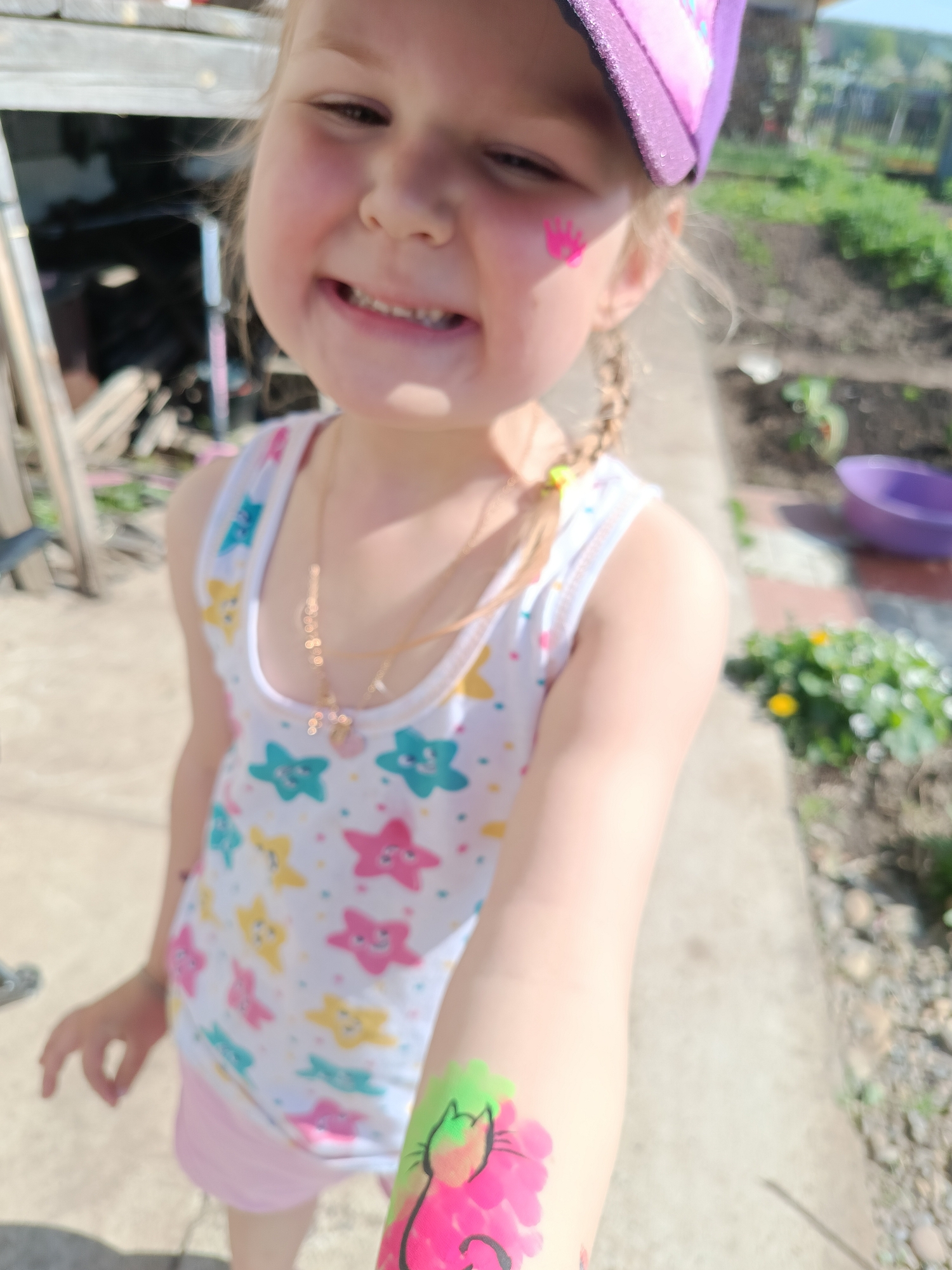 A young girl is smiling at the camera, with a pink crown design painted on her cheek and a pink cat with green ears painted on her arm. She is wearing a white tank top with star patterns, a pink skirt, and a purple baseball cap. She also wears two gold necklaces, one with a cross pendant. She stands outdoors, possibly in a garden, with a sidewalk and various plants and garden implements visible in the background.